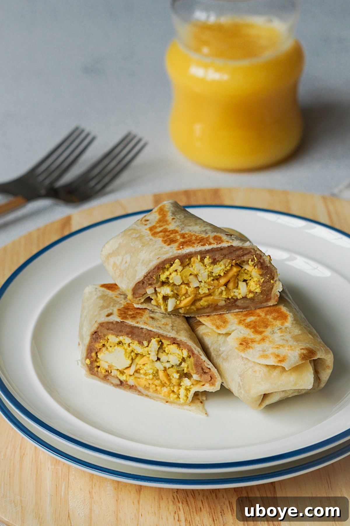 Two vegan breakfast burritos sliced in half on a white plate, served with a glass of orange juice for breakfast