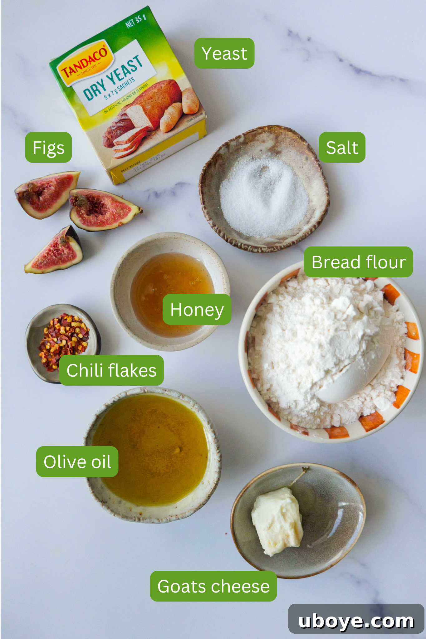 Ingredients for fig and goat cheese focaccia laid out on a marble countertop, including olive oil, goat cheese, chili flakes, flour, honey, fresh figs, salt, and yeast.