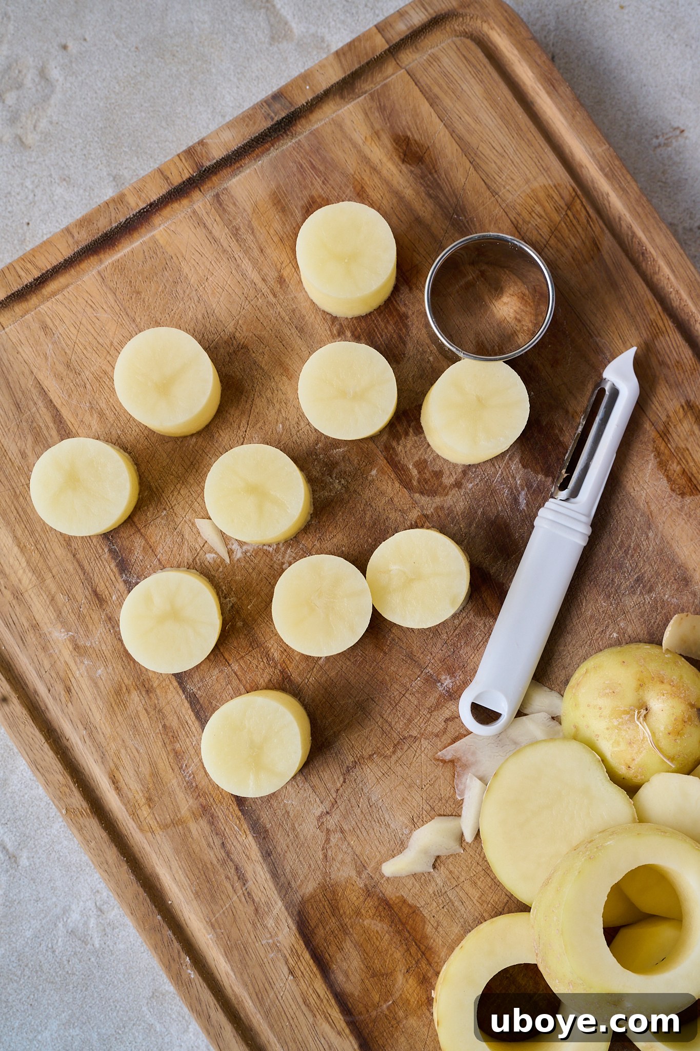 Golden Garlic Thyme Fondant Potatoes 4 Evenly cut potato rounds arranged on a wooden chopping board with a cookie cutter and peeler nearby.