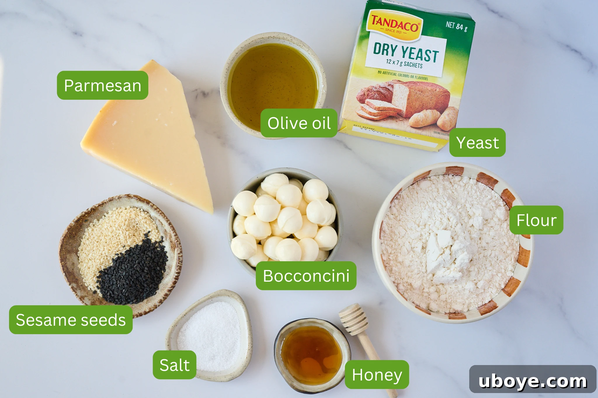 Arrangement of ingredients for cheesy sesame seed focaccia on a marble surface, including parmesan, olive oil, yeast, flour, bocconcini, honey, salt, and sesame seeds.