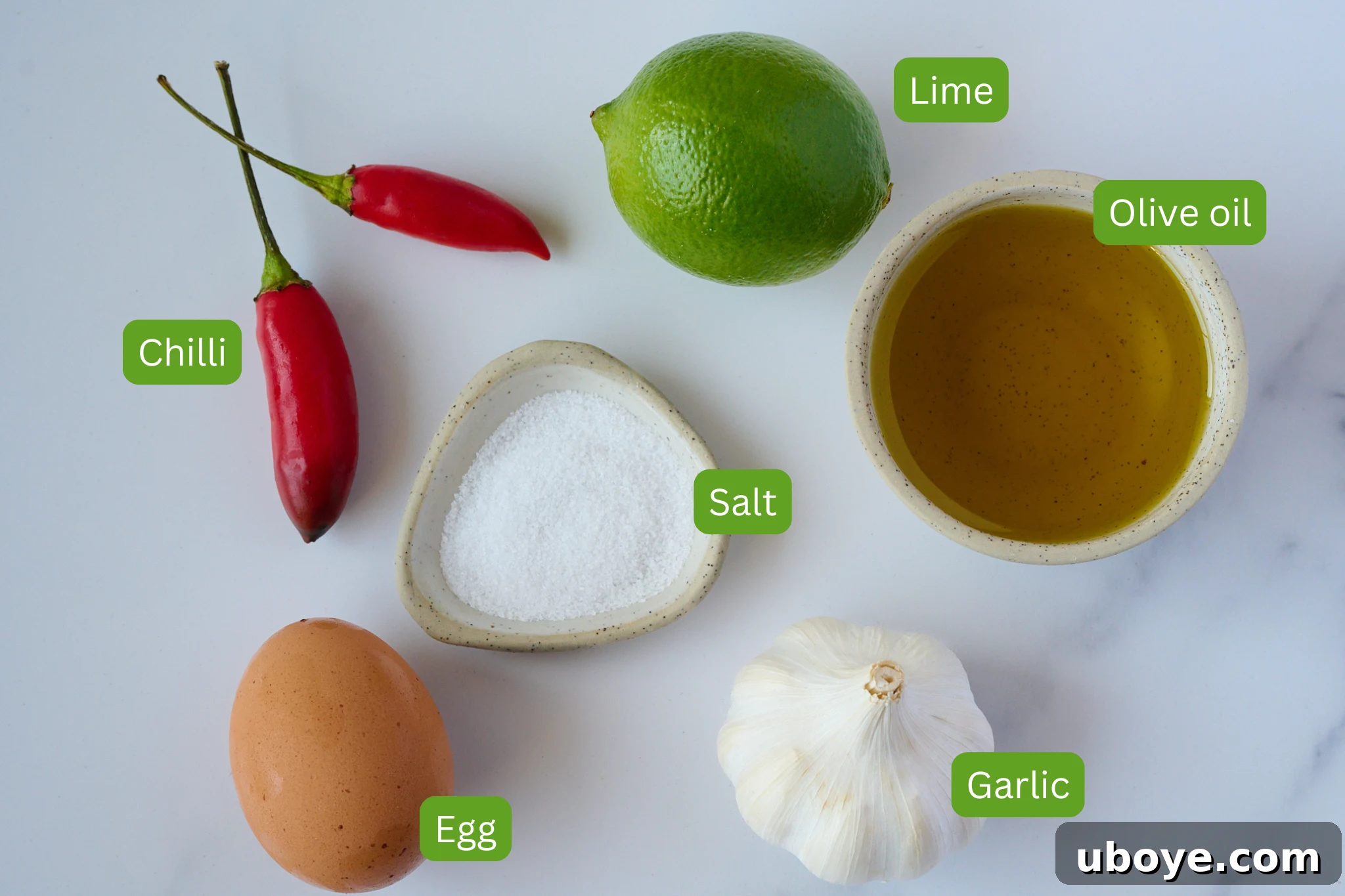 Piquant Chili Garlic Lime Olive Mayonnaise 3 Fresh ingredients for olive oil mayonnaise laid out on a marble background. These include a red chilli, flaky sea salt, a whole lime, a bottle of extra virgin olive oil, a head of garlic, and a single fresh egg.