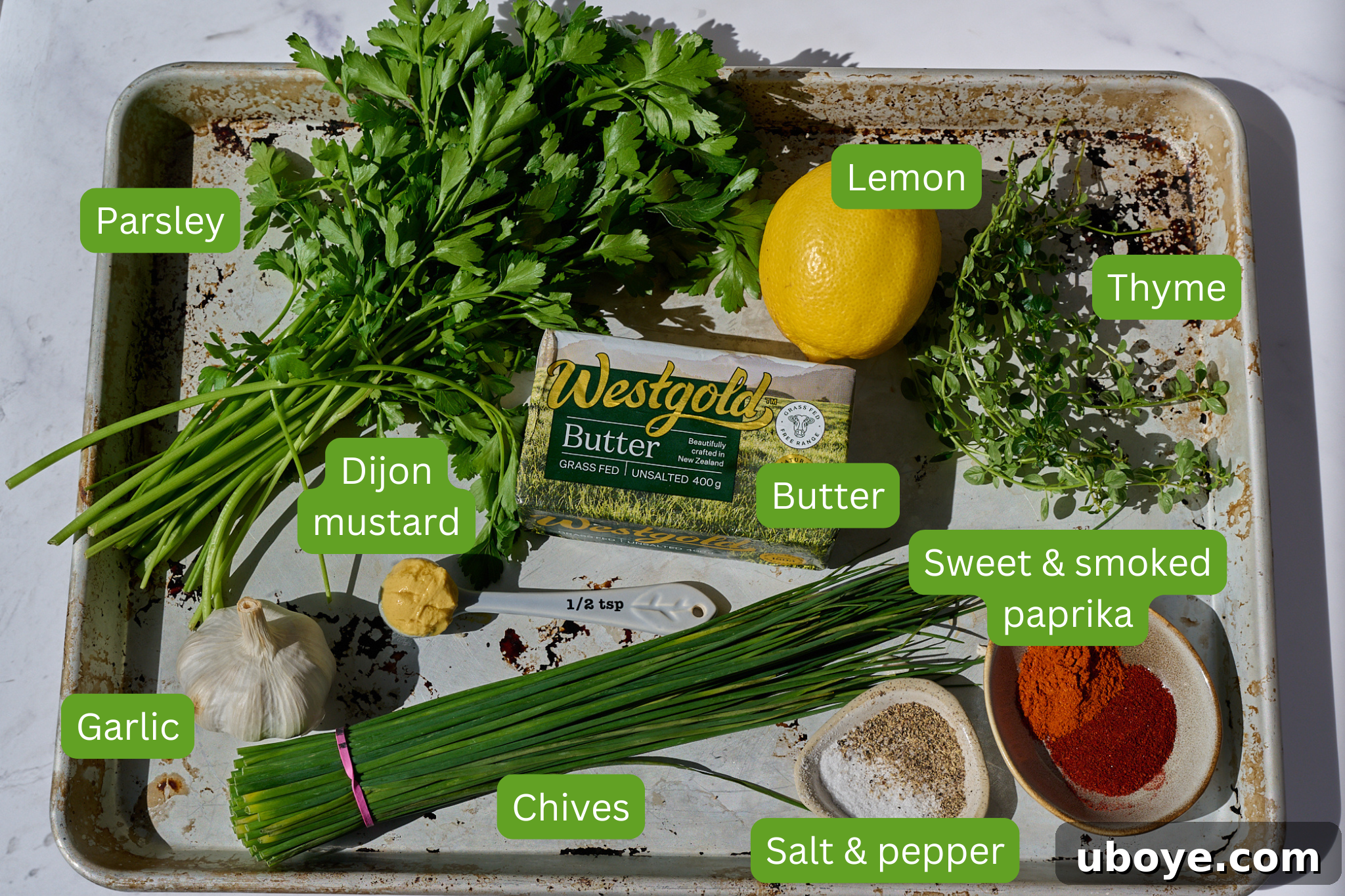 A collection of ingredients for Cowboy Butter, including butter, garlic, fresh herbs, lemon, and various spices, laid out on a wooden board.