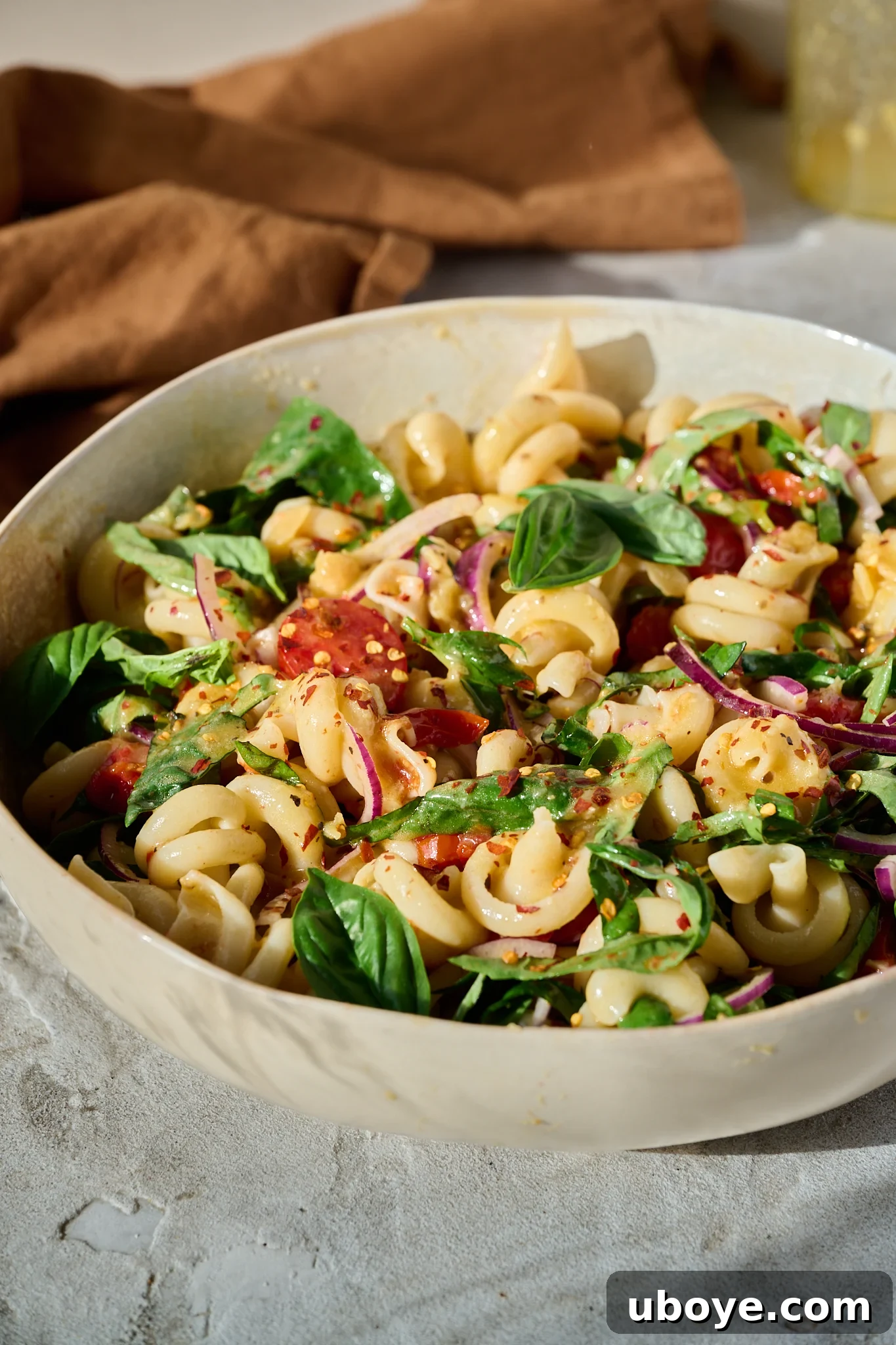 Mediterranean Tomato Confit Pasta Salad sits in large bowl in the sun. There are fresh basil leaves and chilli flakes sprinkled on top.