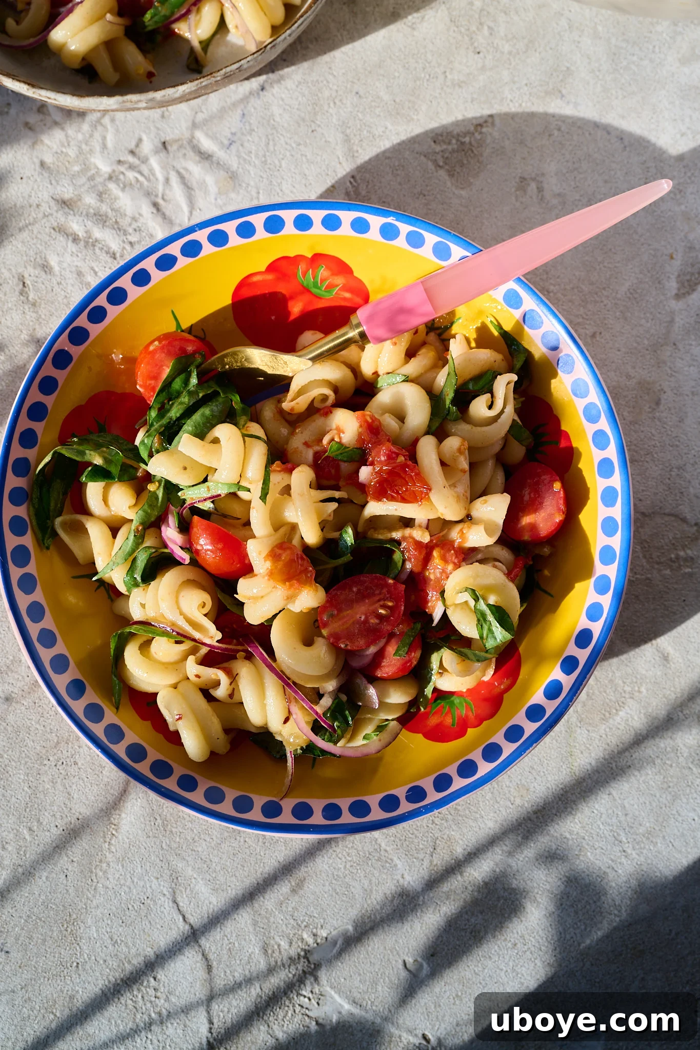Mediterranean Tomato Confit Pasta Salad sits in large bowl in the sun. A pink fork sits out the side of the bowl. The bowl is yellow with a pink and blue rim. There are fresh basil leaves and chilli flakes sprinkled on top.