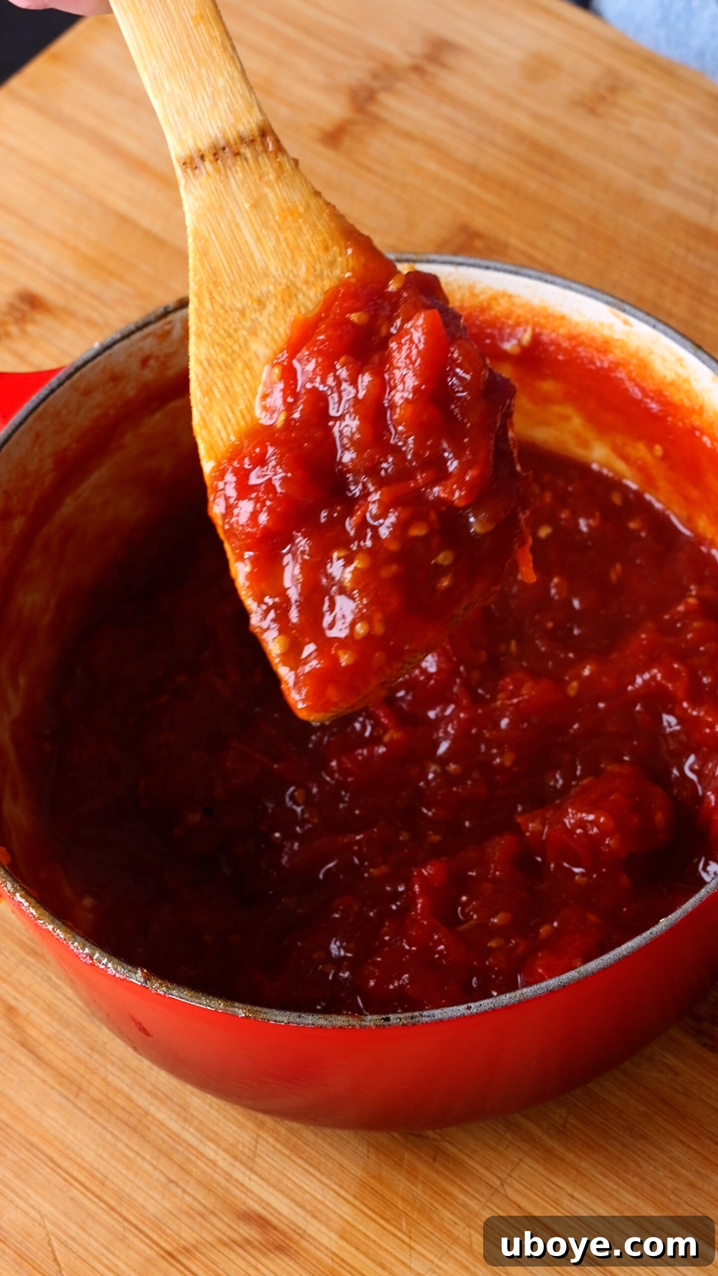 A close-up of a wooden spoon lifting a generous dollop of thick, glossy tomato jam from a red pot, showcasing its rich texture and color after simmering.