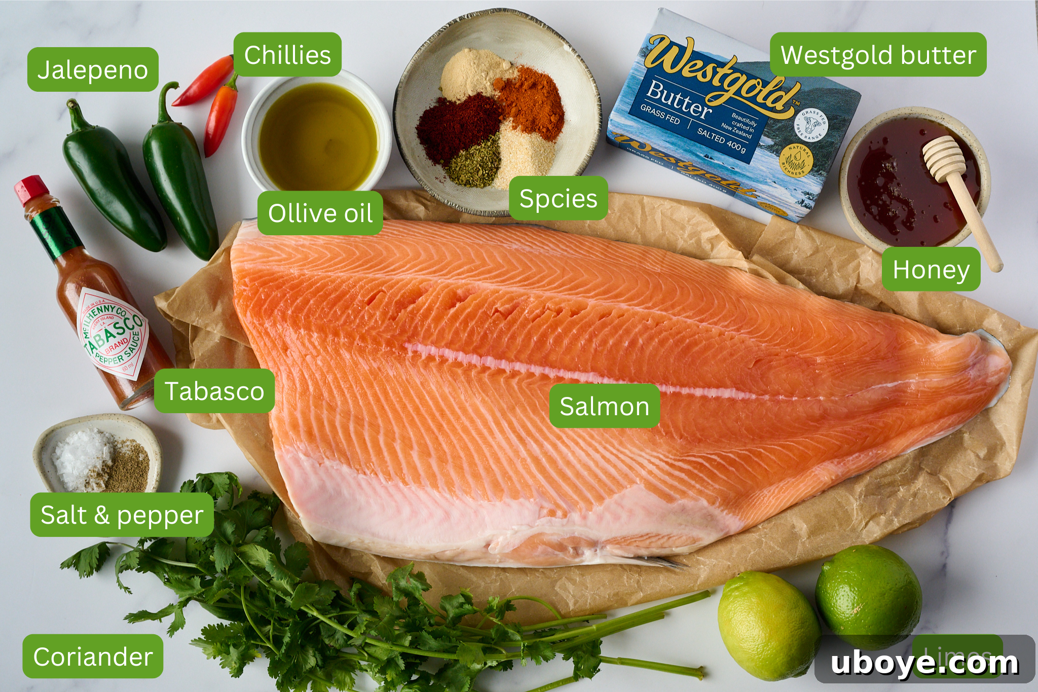 Honey-Chili Slow Roasted Salmon 4 Ingredients for hot honey roasted salmon arranged neatly on a marble counter. Visible items include a large salmon fillet, fresh limes, salt and pepper, vibrant coriander, a bottle of Tabasco sauce, olive oil, jalapeños, various chilies, an assortment of spices, golden honey, and a block of Westgold butter.