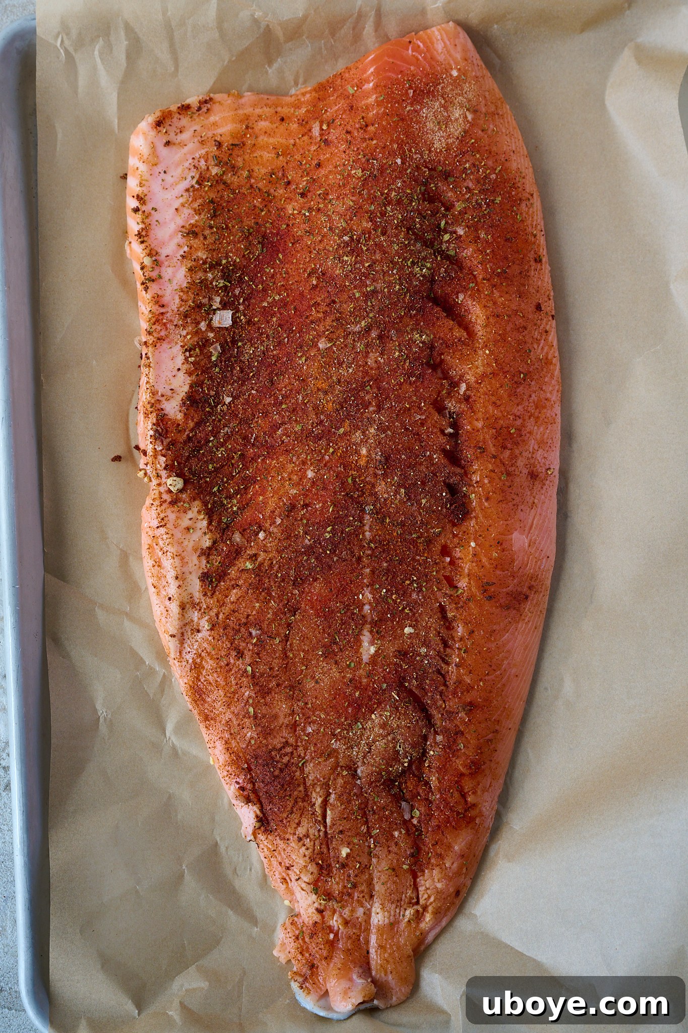 Honey-Chili Slow Roasted Salmon 5 Raw salmon fillet, glistening with a generous coating of various spices, rests on a large baking tray lined with parchment paper, ready for roasting.