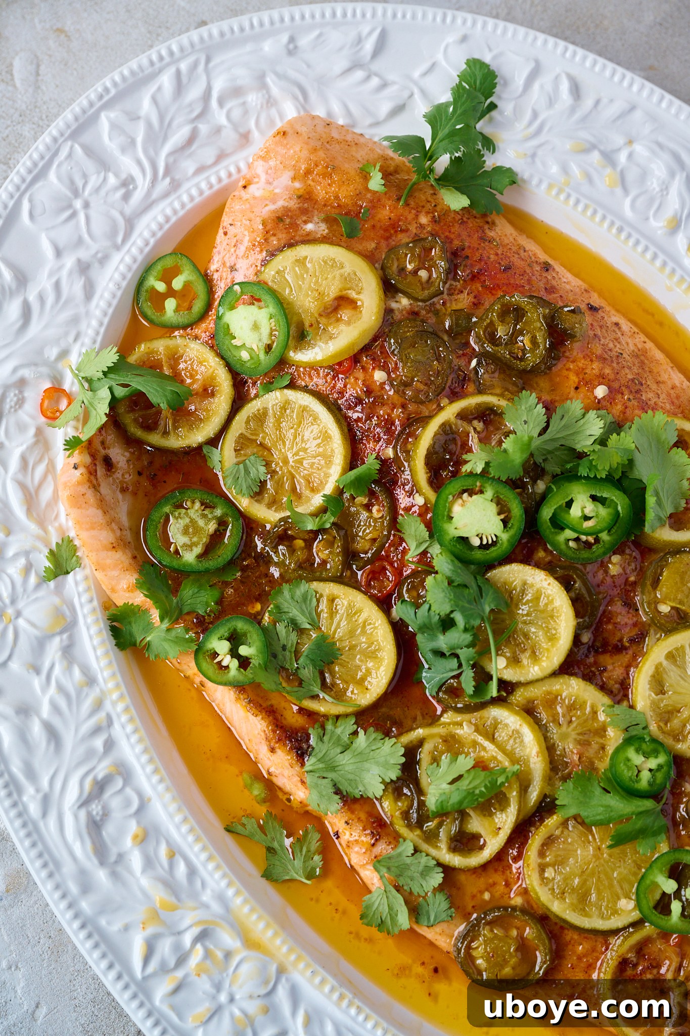 Honey-Chili Slow Roasted Salmon 9 The fully cooked hot honey slow roasted salmon, presented in a large white serving tray, is artfully sprinkled with fresh coriander. The beautifully cooked lime slices and jalapeño peppers are clearly visible, adding to the dish's vibrant appeal.