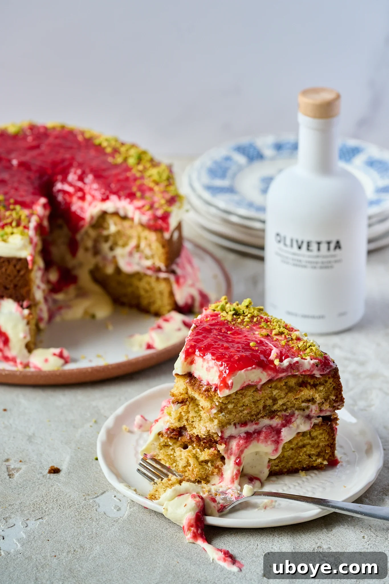 Velvet Olive Oil Cake with Pistachio, Raspberry and White Chocolate 9 A slice of White Chocolate, Pistachio & Raspberry Olive Oil Cake on a white plate with a fork, with the full cake and a bottle of white chocolate olive oil visible in the soft-focus background.
