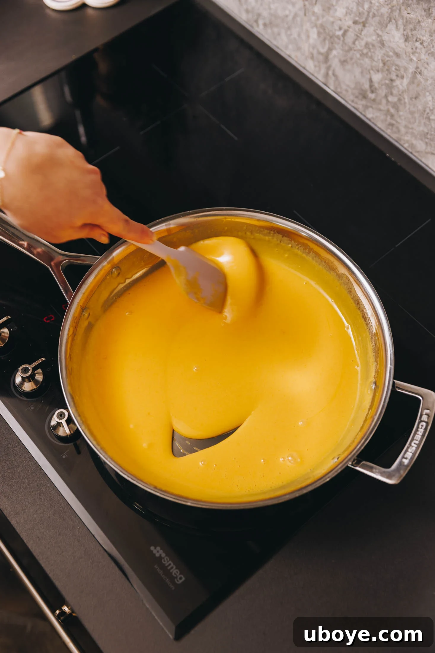 A stainless steel saucepan on a black stovetop, where homemade lemon curd is being gently stirred with a spatula as it thickens.