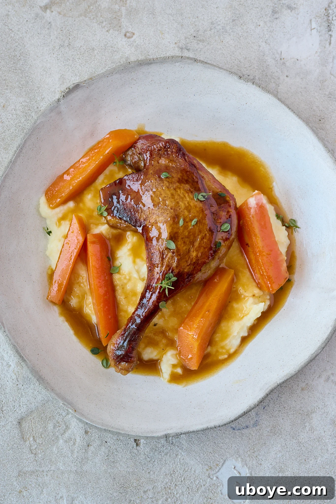 Crispy Braised Duck Legs with Spiced Orange Sauce sits on pile of mash potatoes in large white bowl. Fresh thyme leaves is sprinkled all over. 