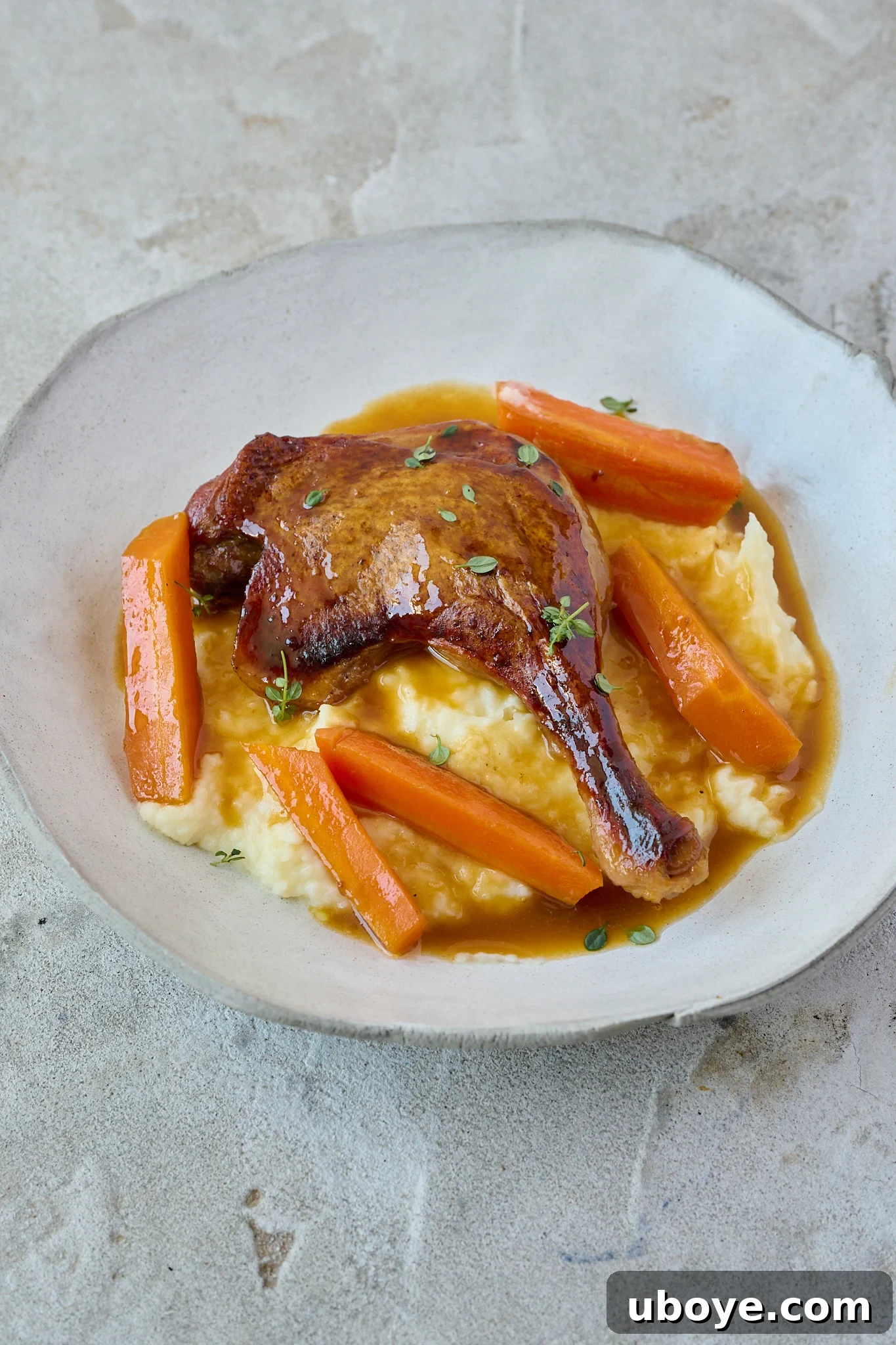 Crispy Braised Duck Legs with Spiced Orange Sauce sits on pile of mash potatoes in large white bowl. Fresh thyme leaves is sprinkled all over. 