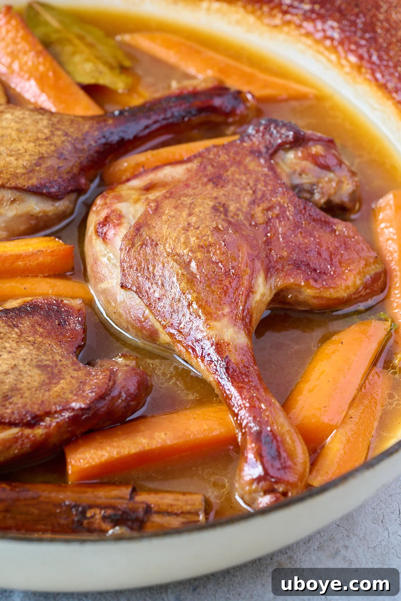 Close up image of crispy duck leg is shown in a white pan with brown sauce and carrots. 