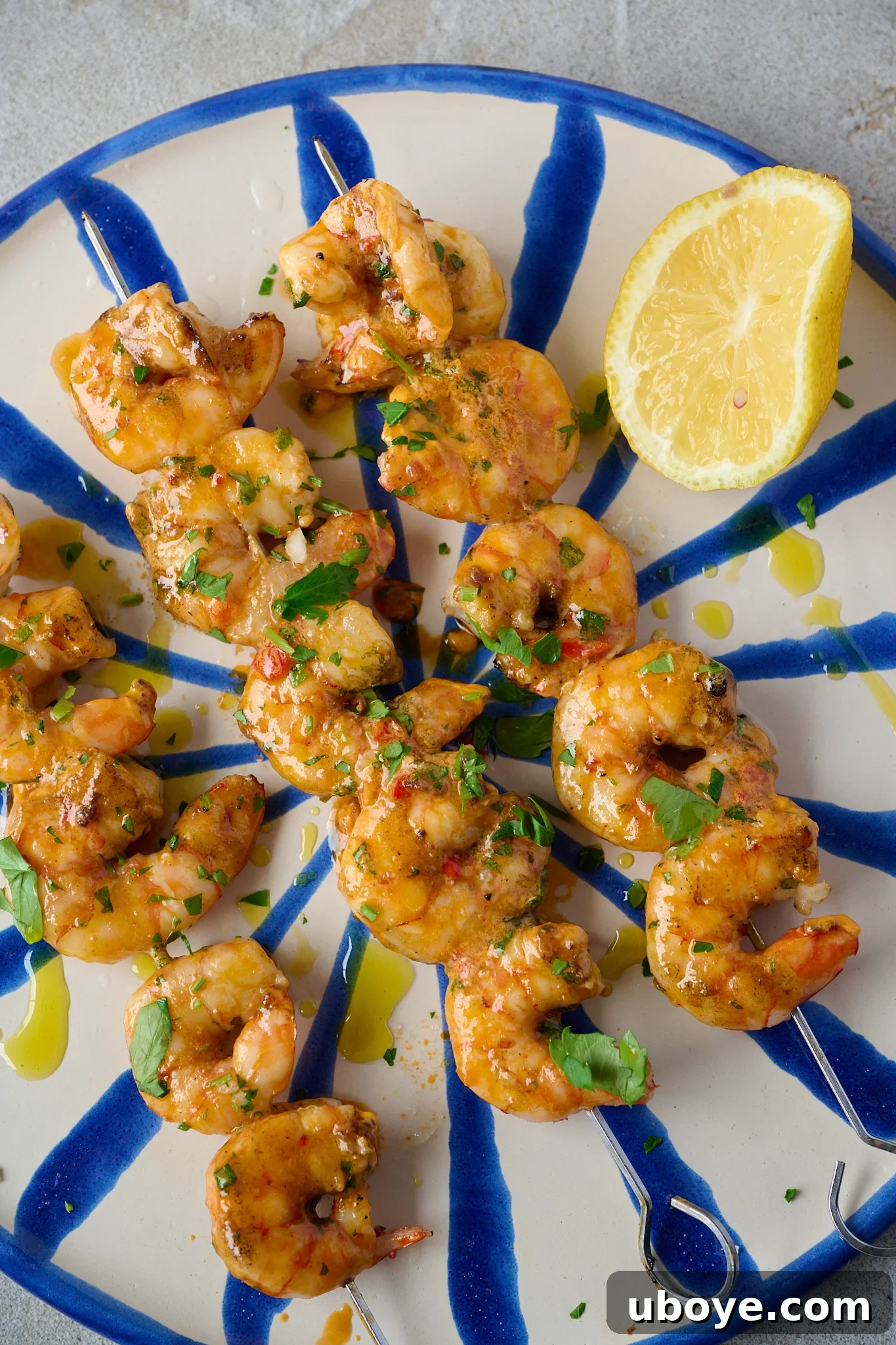 Smoky Grilled Shrimp Skewers with Zesty Tomato Butter 2 Grilled Shrimp Skewers with Tomato Butter (BBQ Prawns) are on blue and white striped plate with fresh parsley sprinkled over. A lemon sits on the right side of the plate.