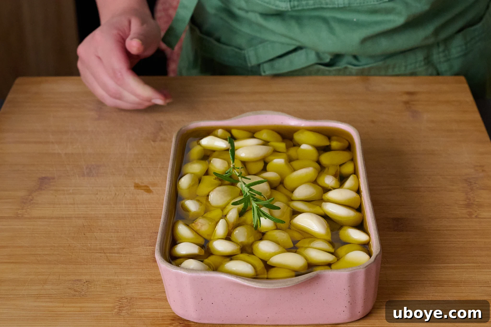Peeled garlic cloves are neatly arranged in a pink baking dish, completely submerged in golden olive oil, adorned with a fragrant sprig of fresh rosemary.