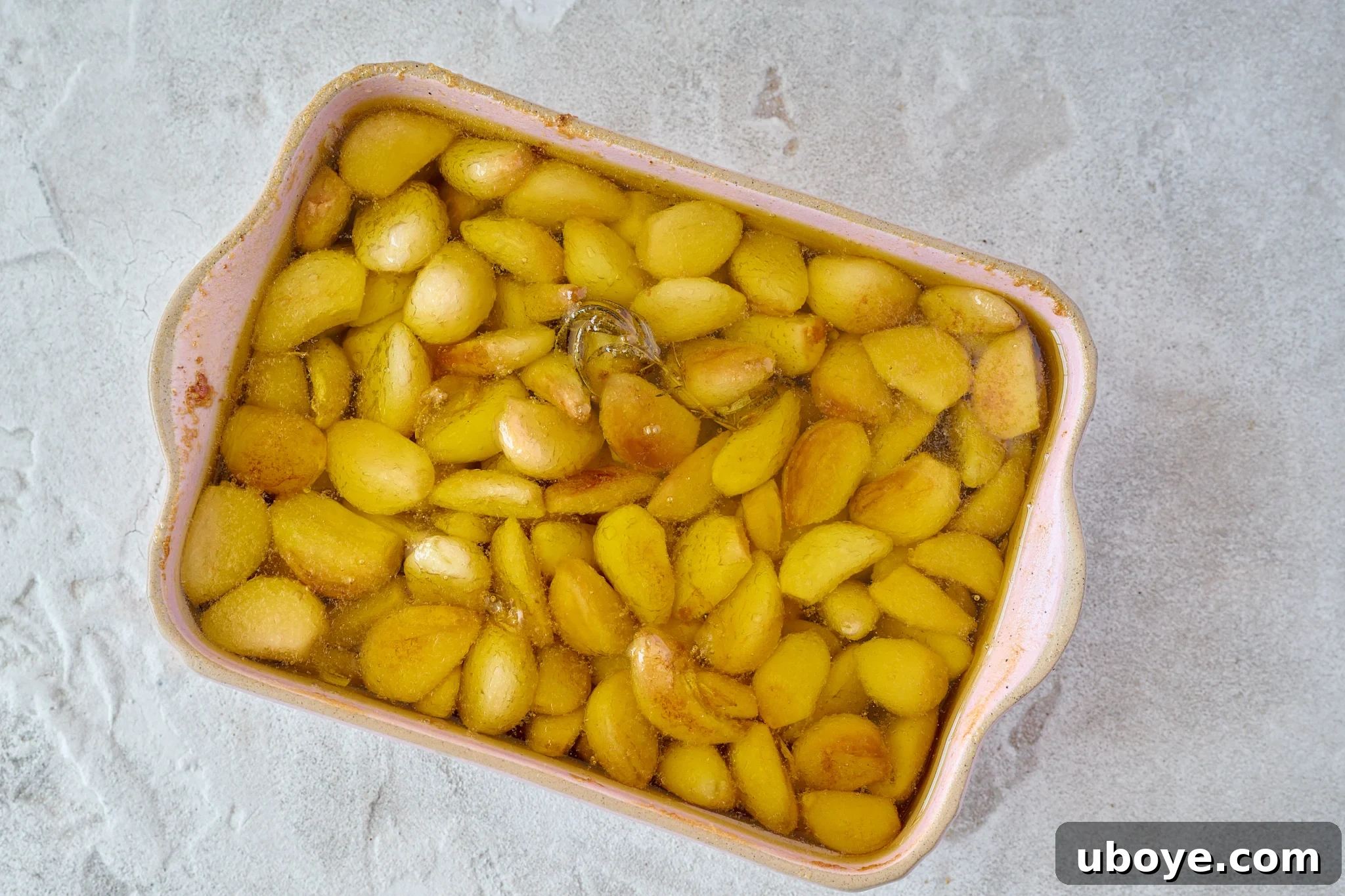Perfectly golden brown, tender oven-roasted garlic confit cloves are visible in a pink baking dish, softened and beautifully infused in the olive oil.