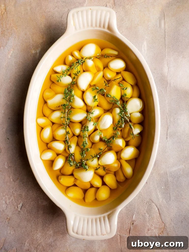 Fresh raw garlic cloves arranged in a white baking dish, fully submerged in clear olive oil, accompanied by a sprig of aromatic rosemary, ready for the oven.