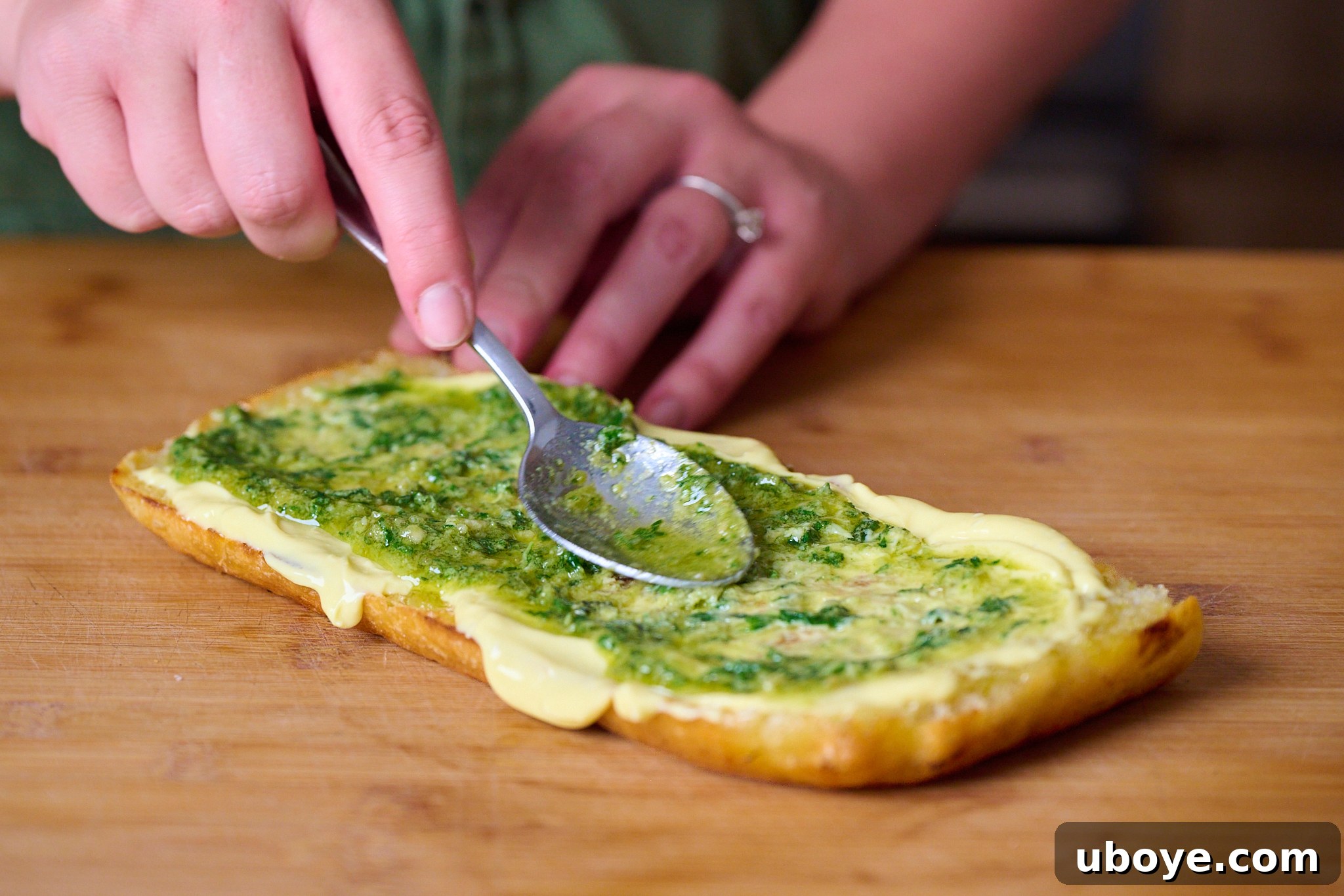 Piece of focaccia that has been slightly toasted and now has a layer of garlic confit aioli and pesto. Sits on wooden chopping board. 