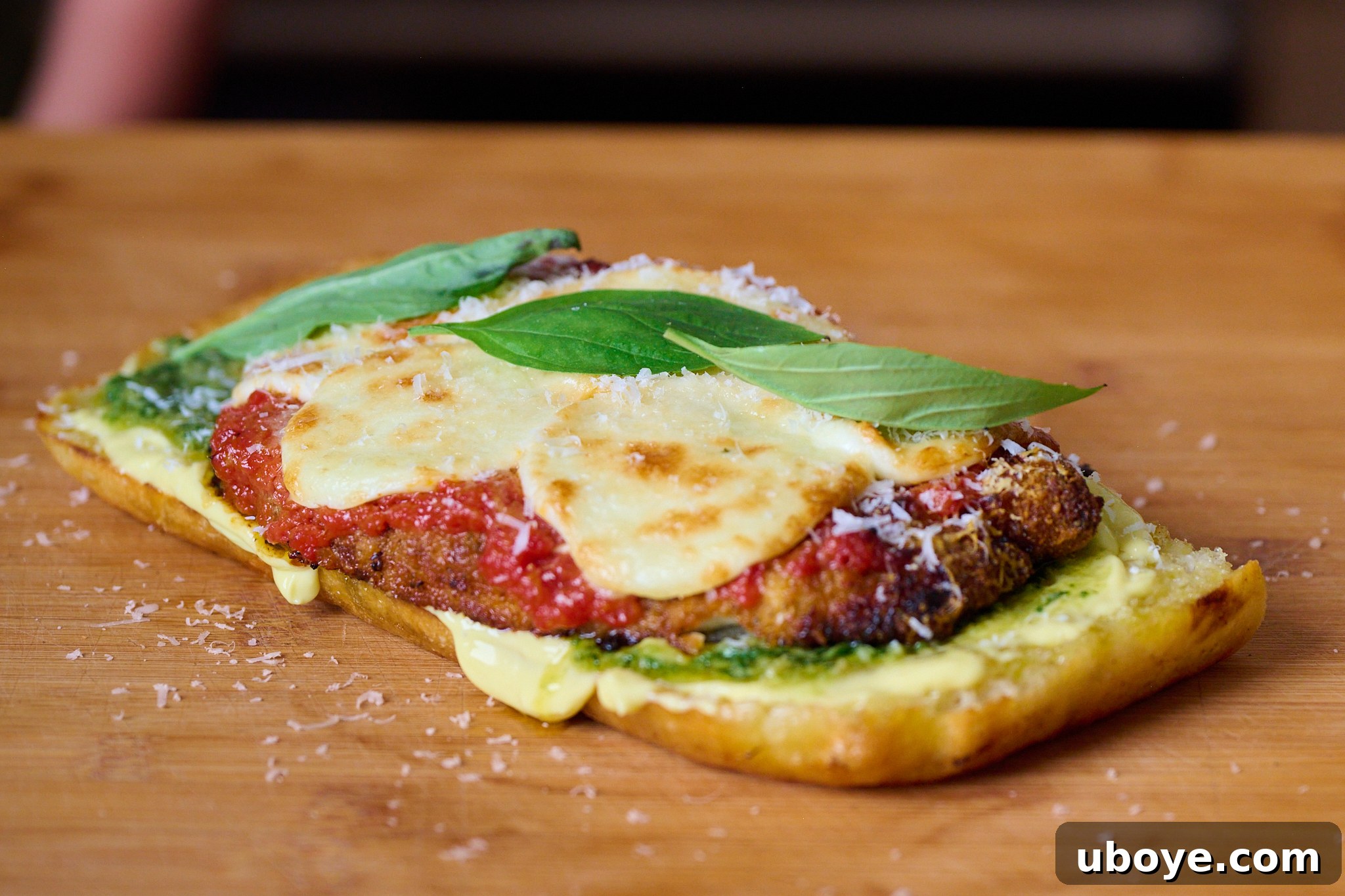 Piece of focaccia that has been slightly toasted and now has a layer of garlic confit aioli, pesto and the chicken parm being put on top. Freshly grated parmesan and basil leaves sit on top of chicken parma. Sits on wooden chopping board. 