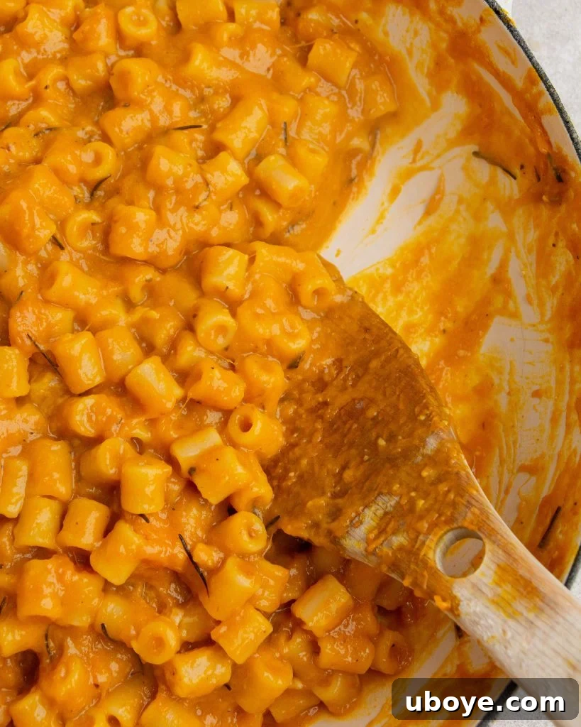 A large white pot is filled with roasted pumpkin and garlic pasta, with a wooden spoon casually resting inside on the right side. 