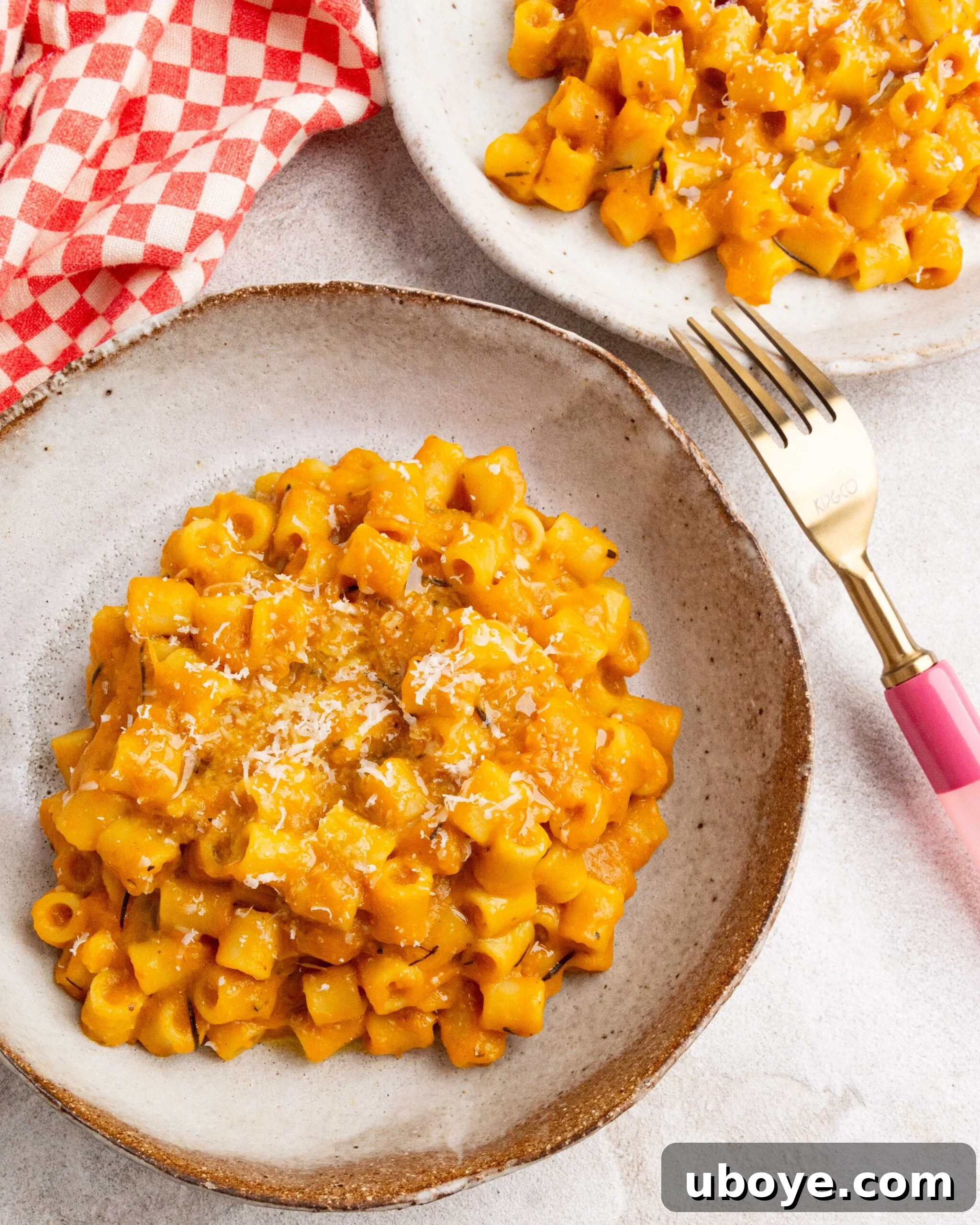 A brown bowl holds roasted pumpkin and garlic pasta, with a pink fork on the right. In the background, a red and white checkered tea towel and another bowl of pasta are visible.