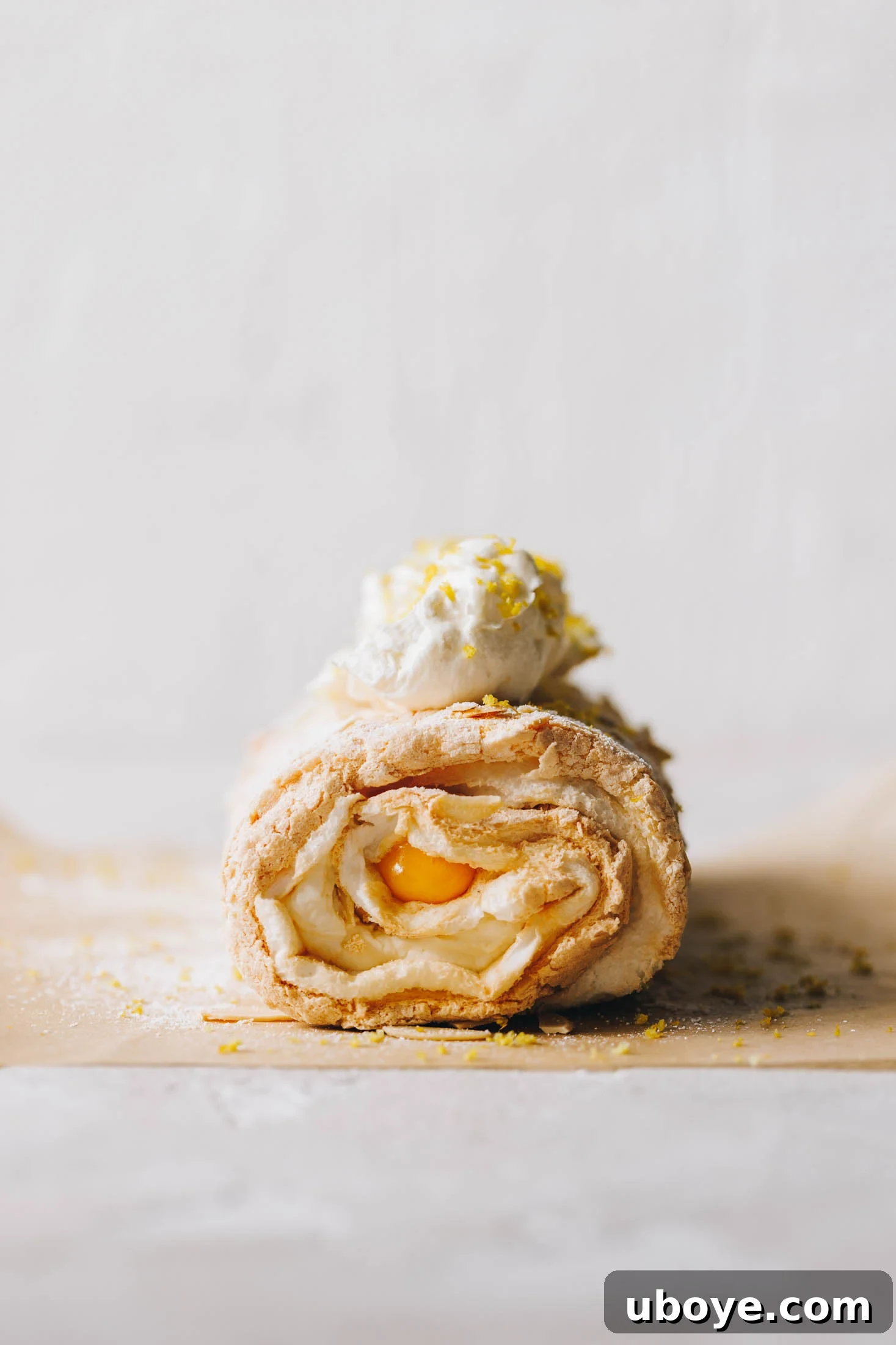 Lemon curd meringue roulade sits on baking paper and has whipped cream and lemon zest on top. 