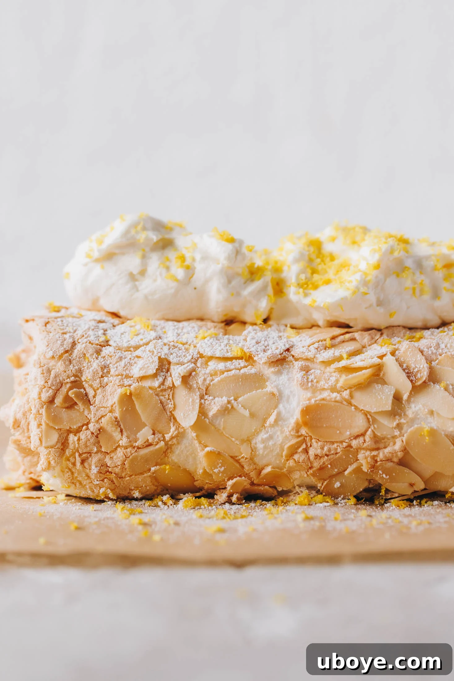Lemon curd meringue roulade sits on baking paper and has whipped cream and lemon zest on top. 