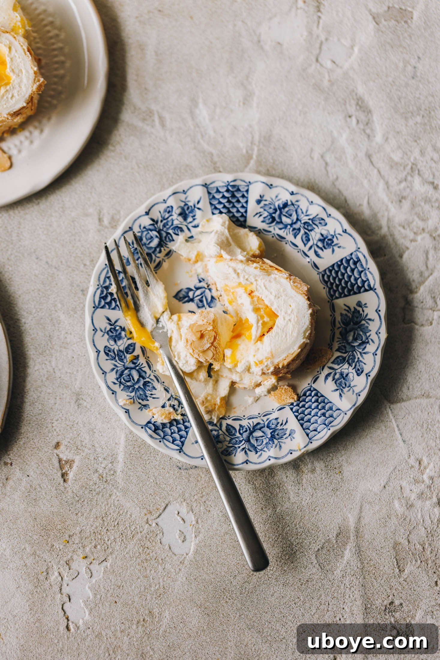 A slice of lemon curd meringue roulade sits on a blue and white plate with a floral pattern. A slice has been taken out of it with a fork. 