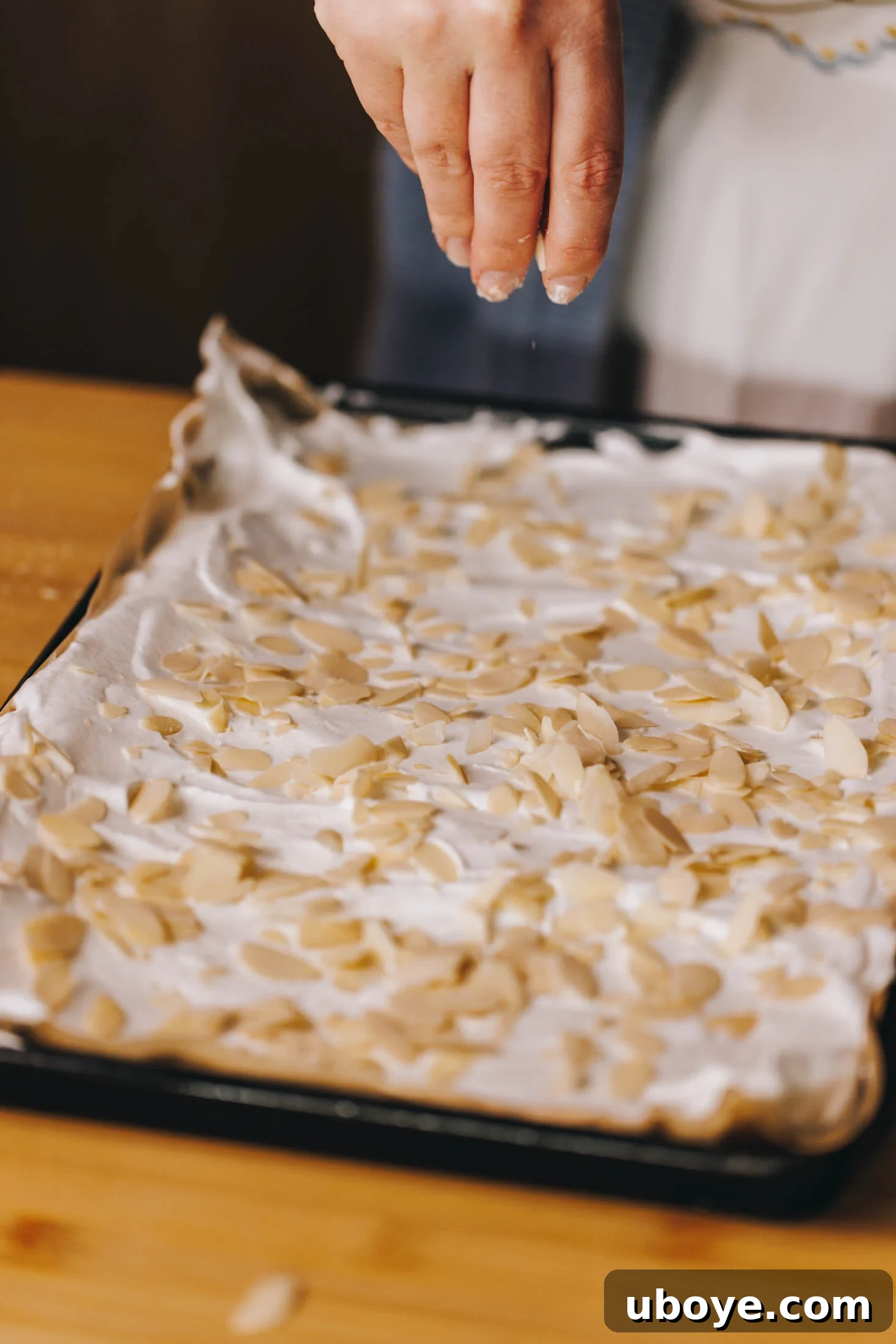 Slithered almonds are being sprinkled onto meringue on baking tray. 