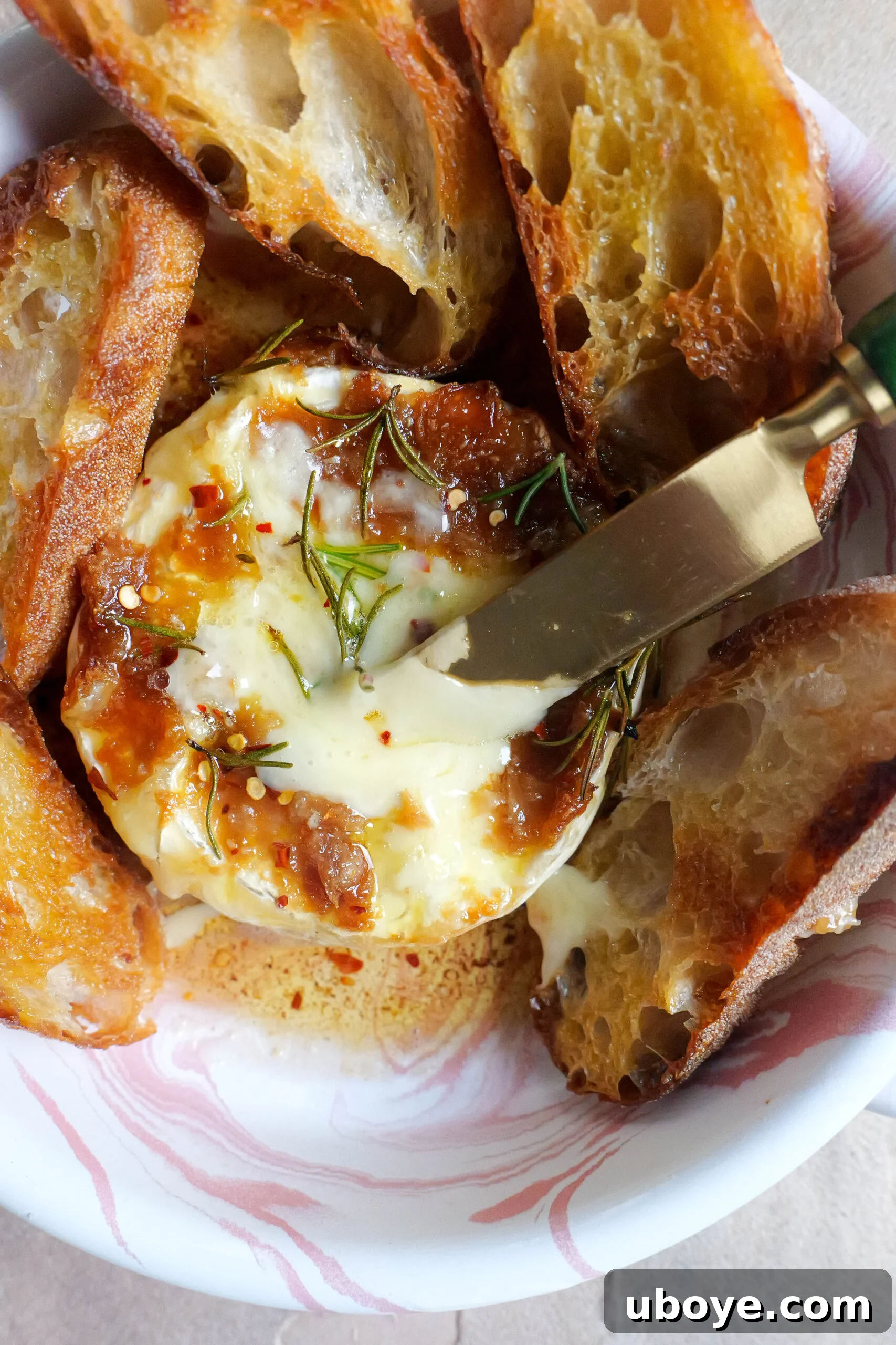 Holiday Roasted Garlic Butter Baked Brie 5 Baked brie with roasted garlic butter sits in pink and white tray with olive oil fried slices of baguette. There is also a knife sticking out of the brie.
