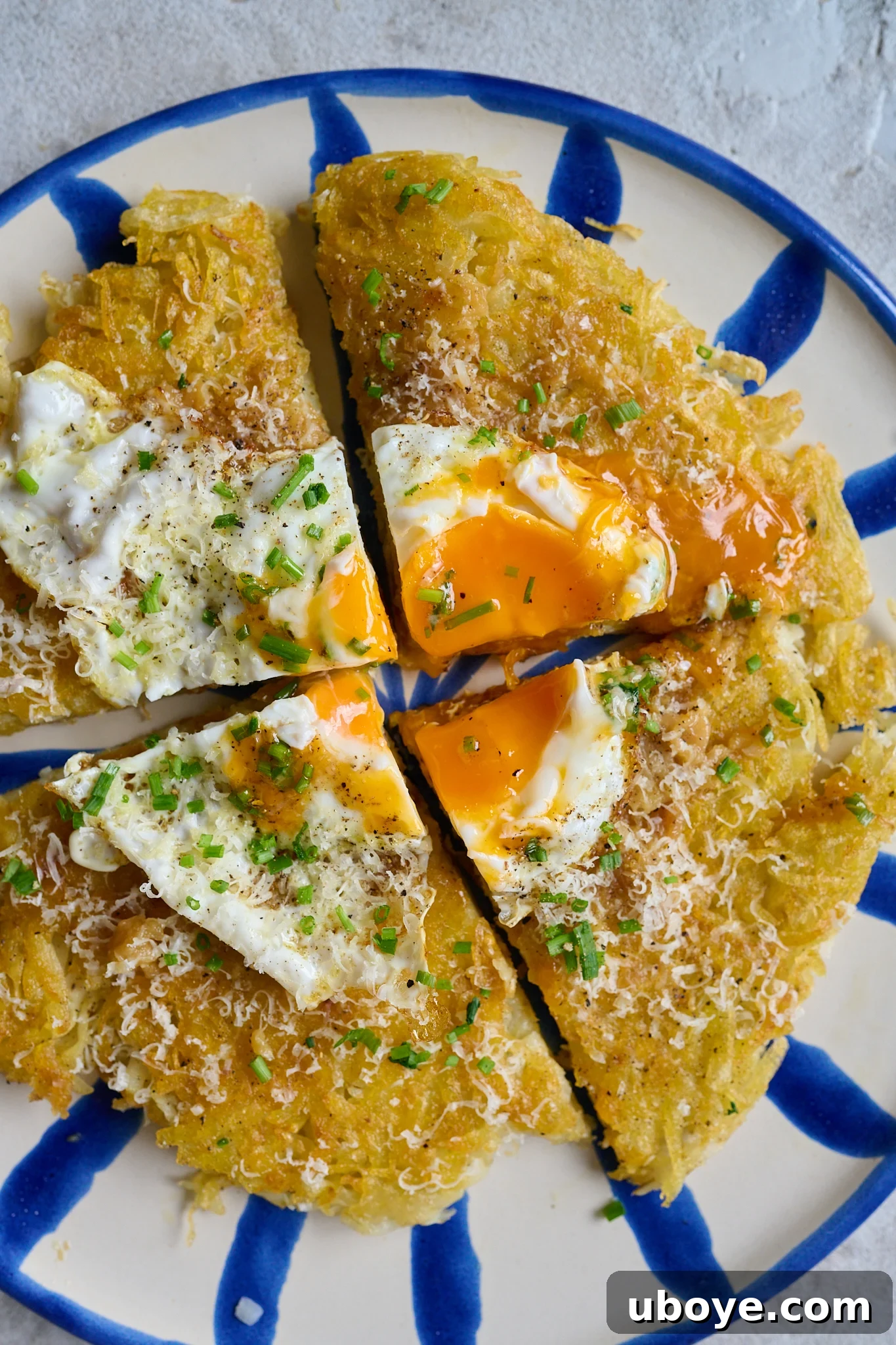 Golden Crispy Potato Rosti 11 A large potato rosti cut into quarters, topped with a fried egg whose yolk has broken, sprinkled with chives and cheese, served on a white plate with a blue rim.