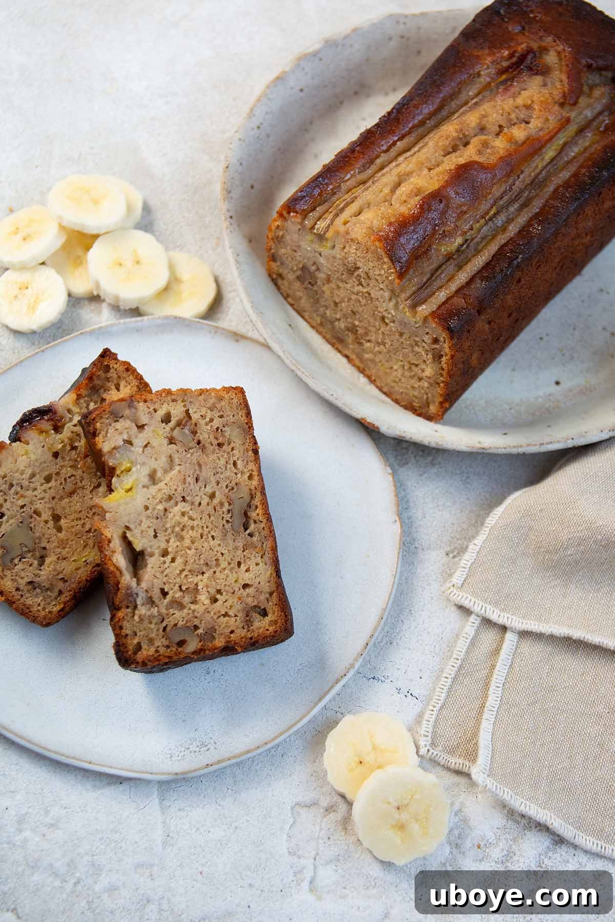 Two slices of fluffy olive oil banana bread on a white plate with the full loaf in the background, beautifully baked.