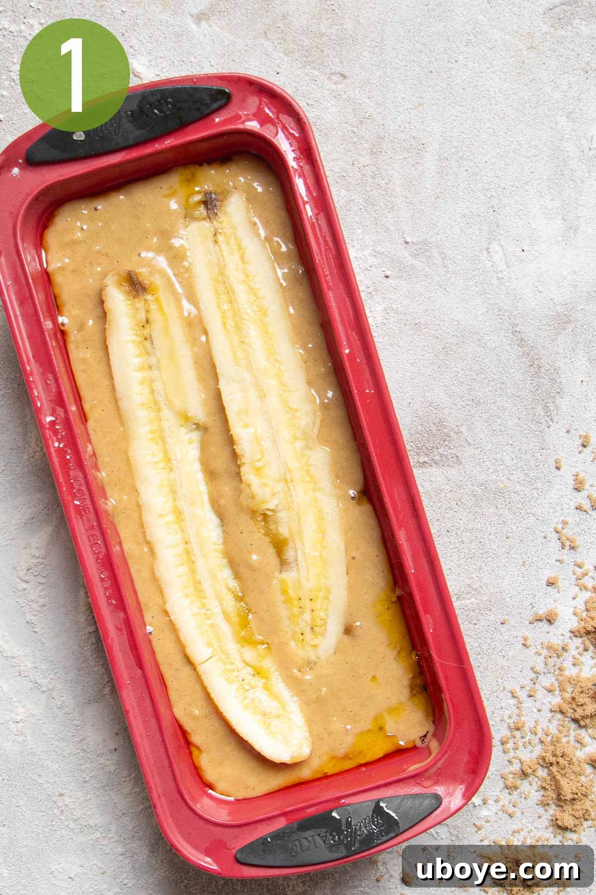 Unbaked banana bread batter in a vibrant red loaf pan, topped with a halved banana, ready for the oven.