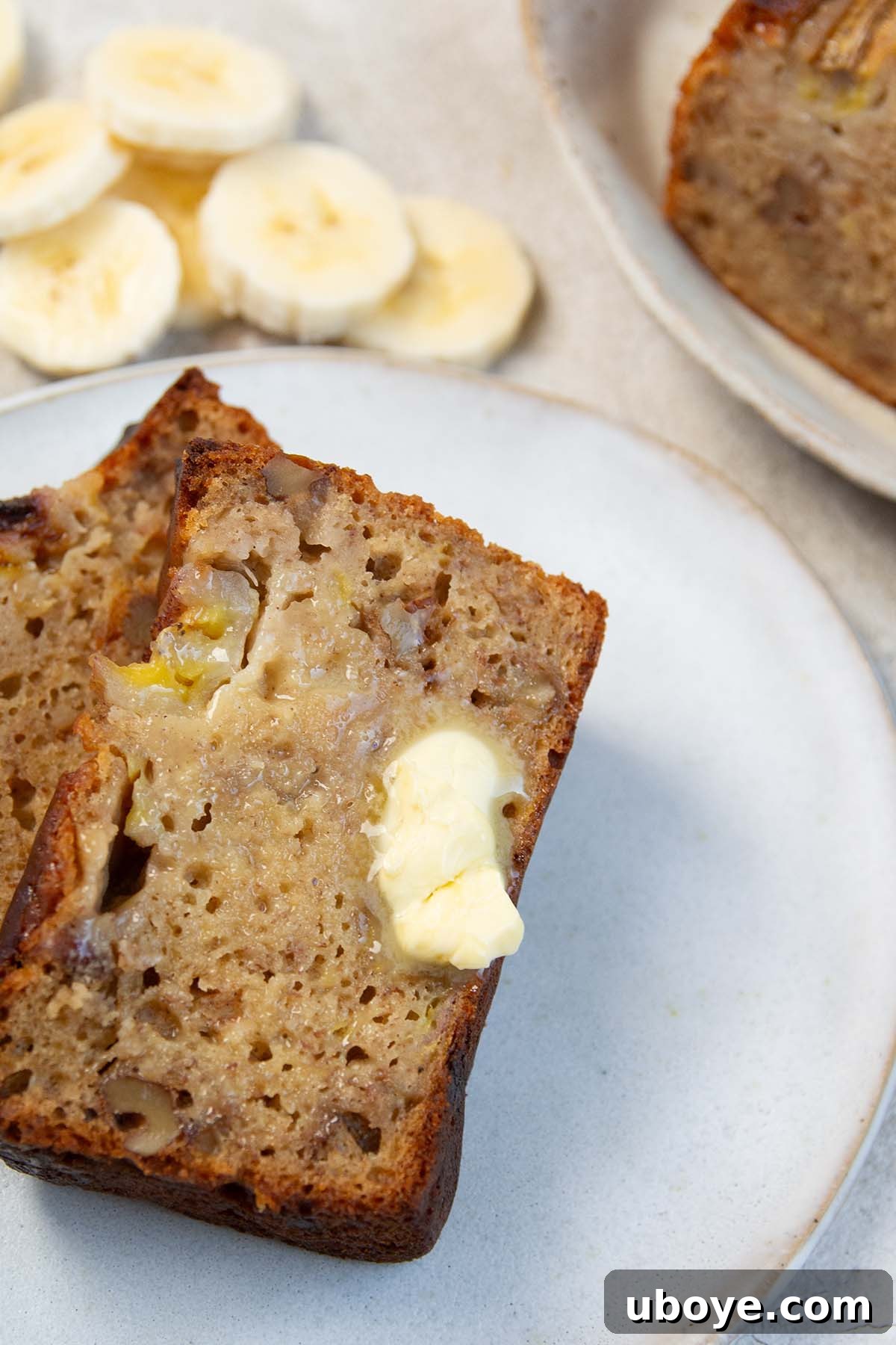 A slice of olive oil banana bread, generously buttered, resting on a pristine white plate, inviting a delicious bite.