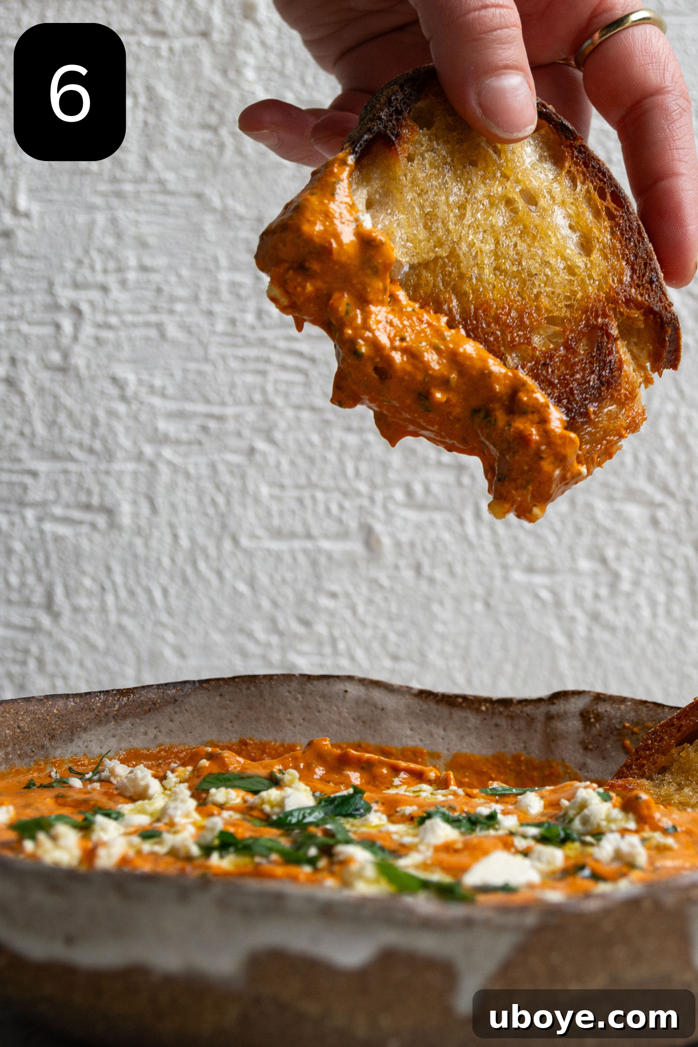 Fiery Feta and Red Pepper Dip 9 A hand holding a piece of golden, crispy sourdough bread, dipping it into the vibrant red pepper and feta dip, ready to be enjoyed.