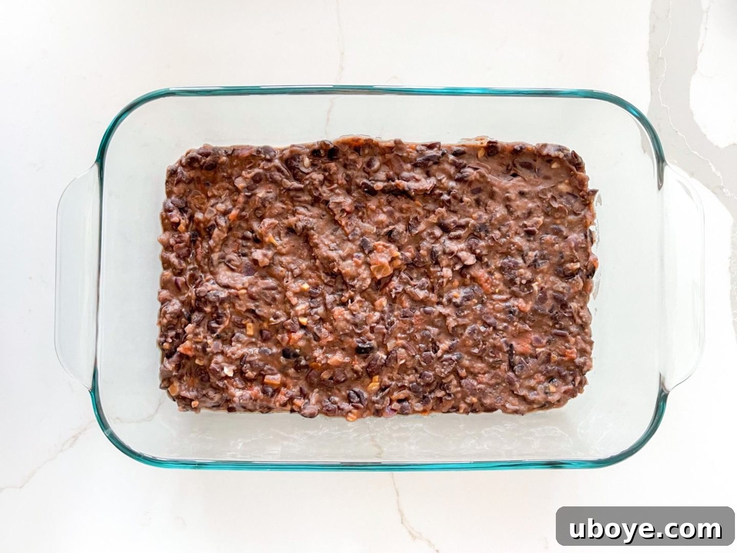 Epic Vegan 7 Layer Dip 6 Smooth, dark refried black beans spread evenly in a glass casserole dish, cooling before the next layer is added.