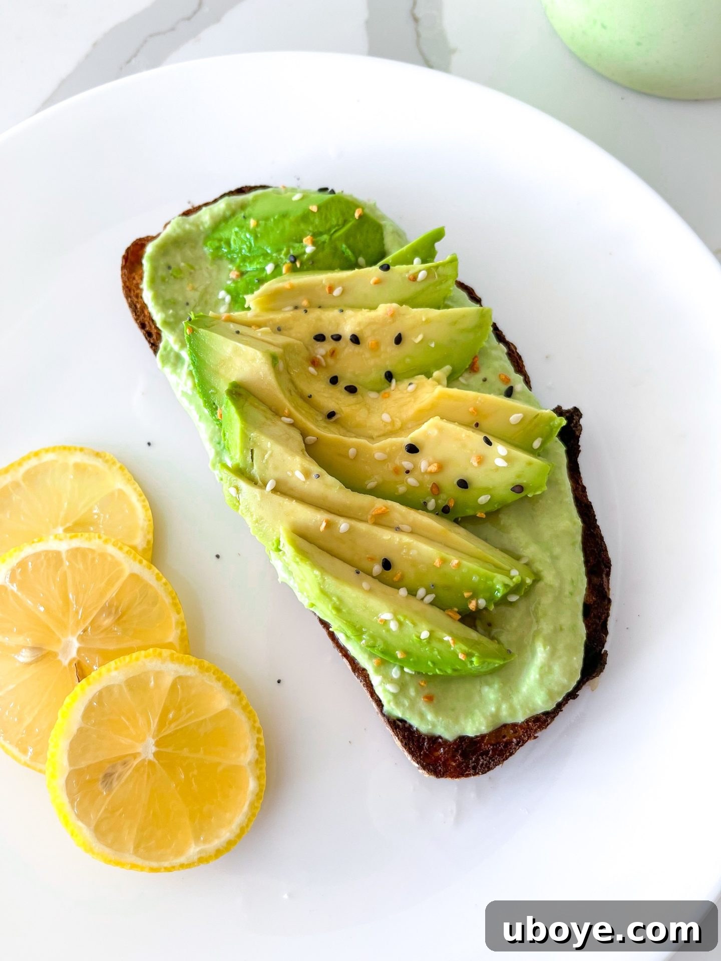 Bright Lemon Lima Hummus 6 A delectable piece of sourdough toast generously topped with creamy lima bean hummus and fresh avocado slices, presented on a pristine white plate.