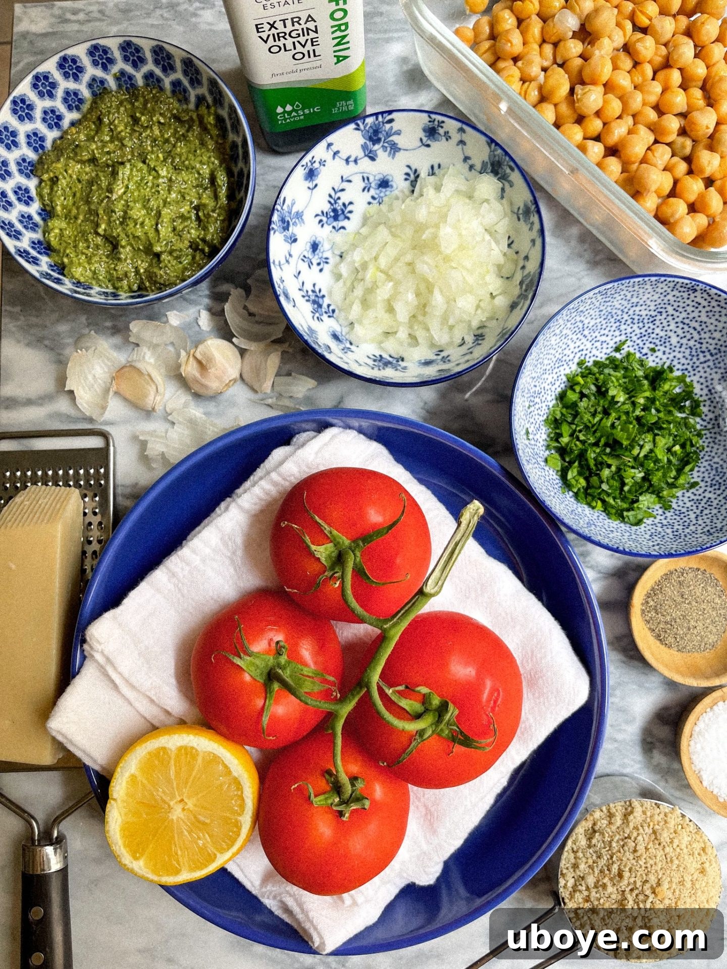 Pesto-Packed Tomato Delights 4 Fresh ingredients laid out on a kitchen counter: whole tomatoes, red onion, a bowl of vegan pesto, a block of vegan Parmesan, and fresh herbs.