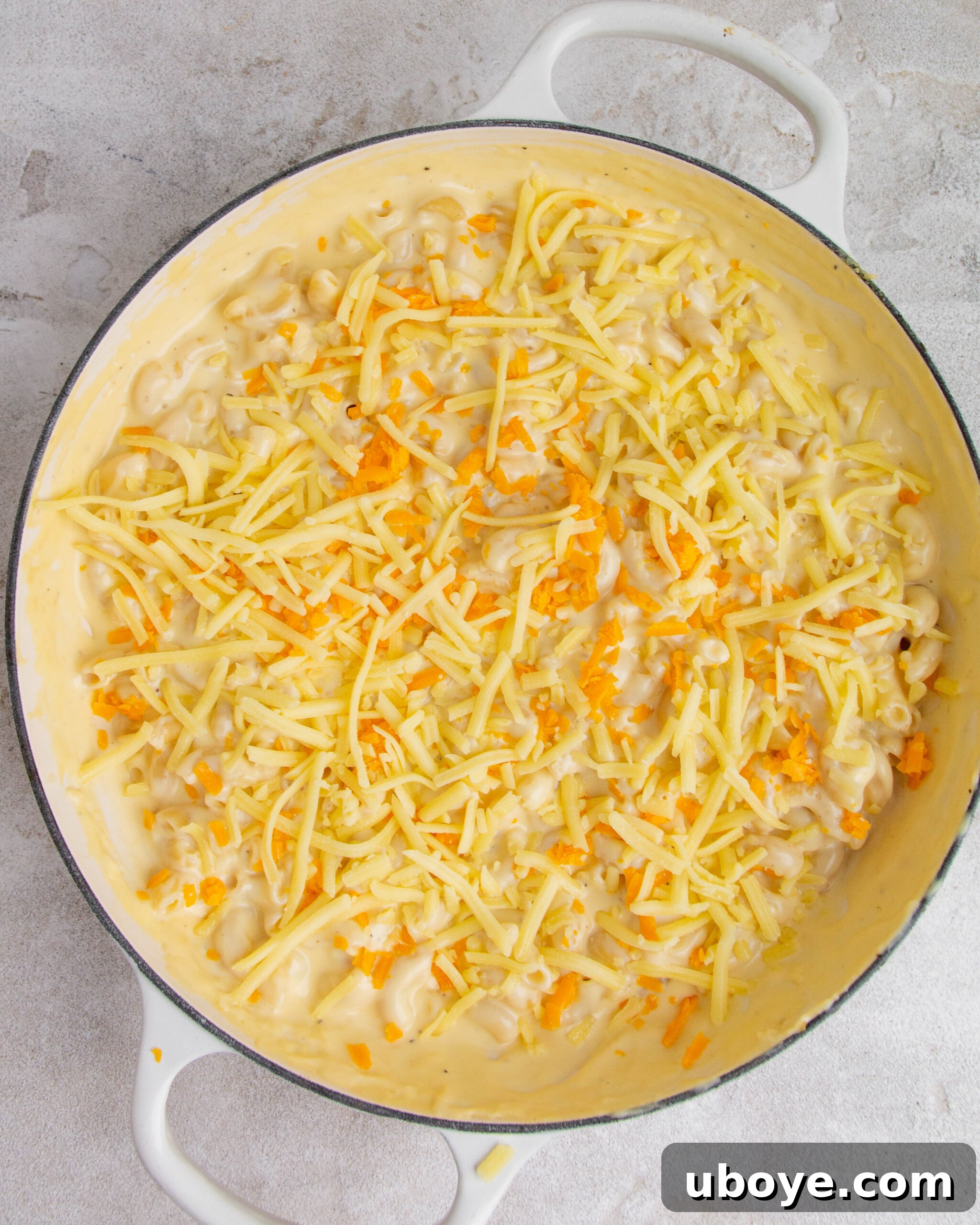 Unbaked mac and cheese with garlic confit in a large white baking tray, topped with a generous layer of shredded cheese.