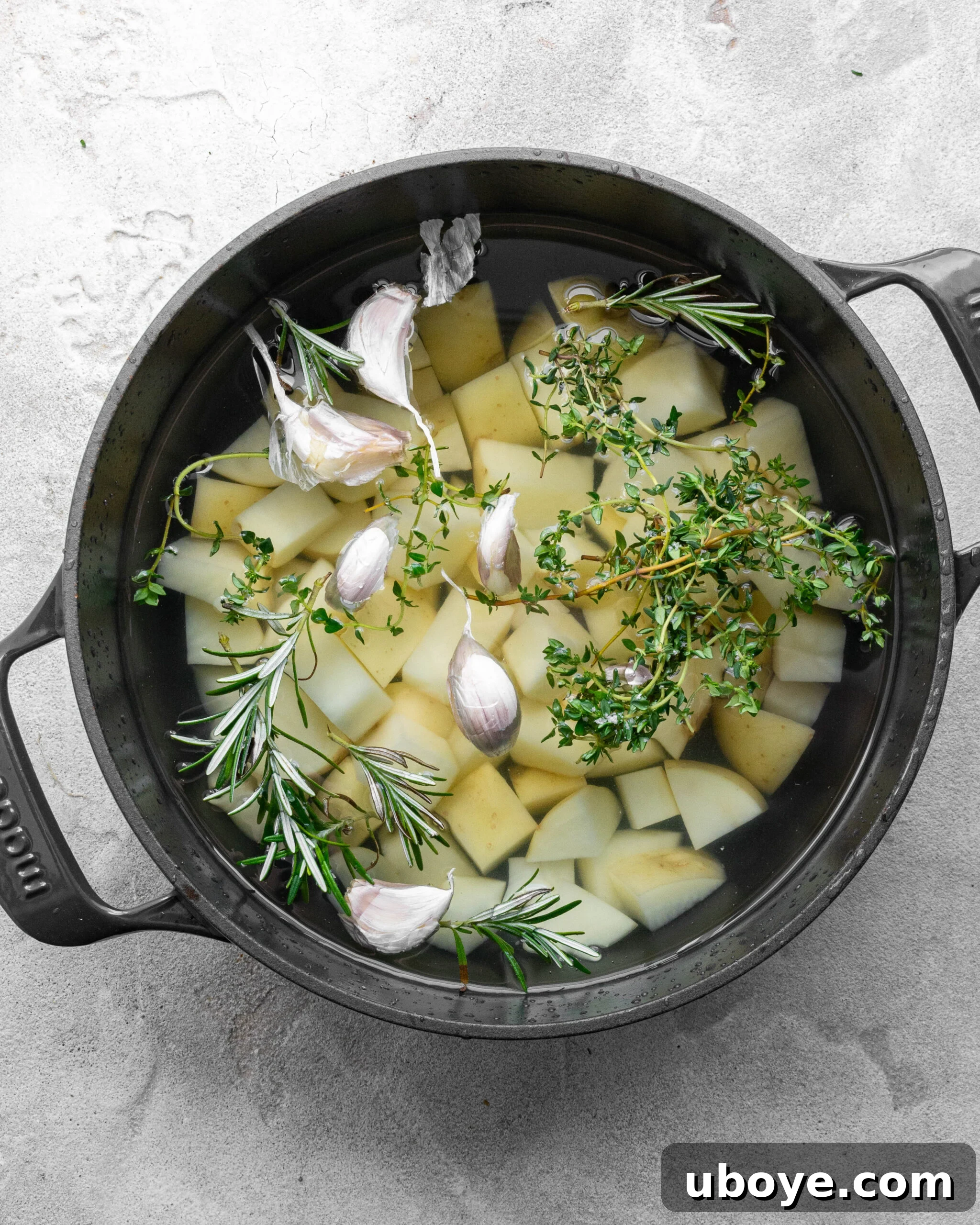 Peeled and cut potatoes soaking in cold water in a large blue pot with fresh garlic, sprigs of thyme, and rosemary, ready for the boiling process.
