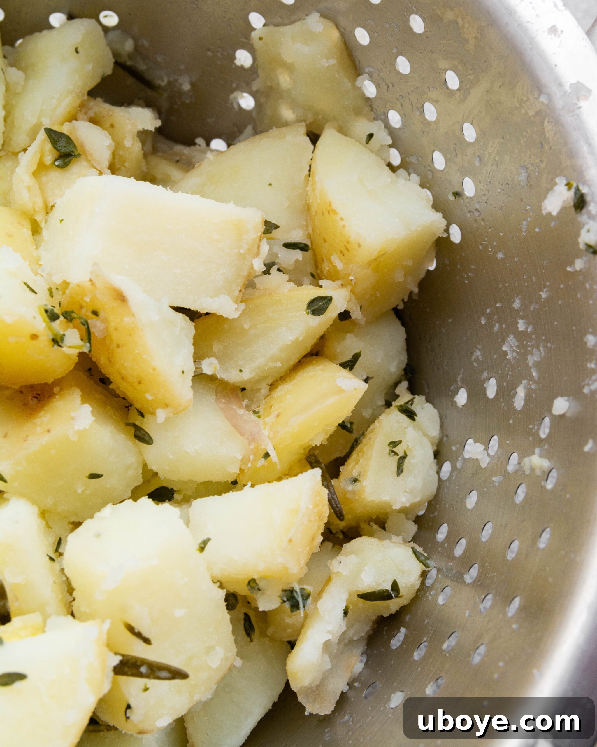 Cooked and roughed-up potatoes resting in a stainless steel colander, covered with a tea towel to steam-dry.