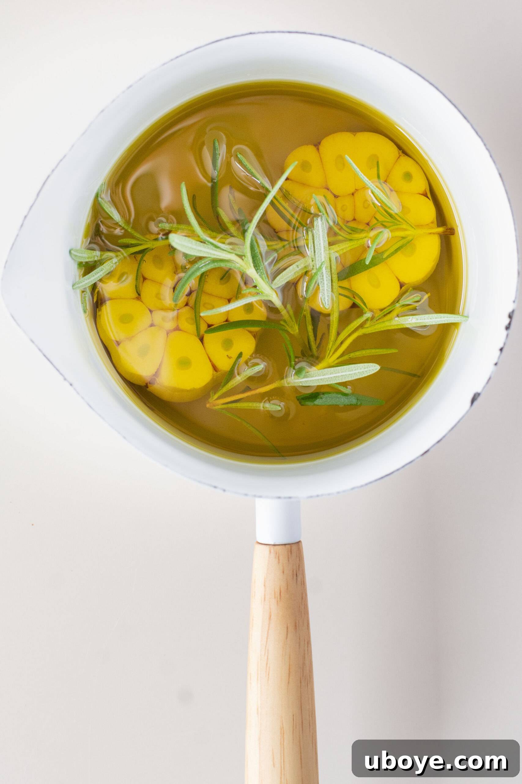 Small white pot with olive oil, raw garlic bulbs, and a sprig of rosemary.