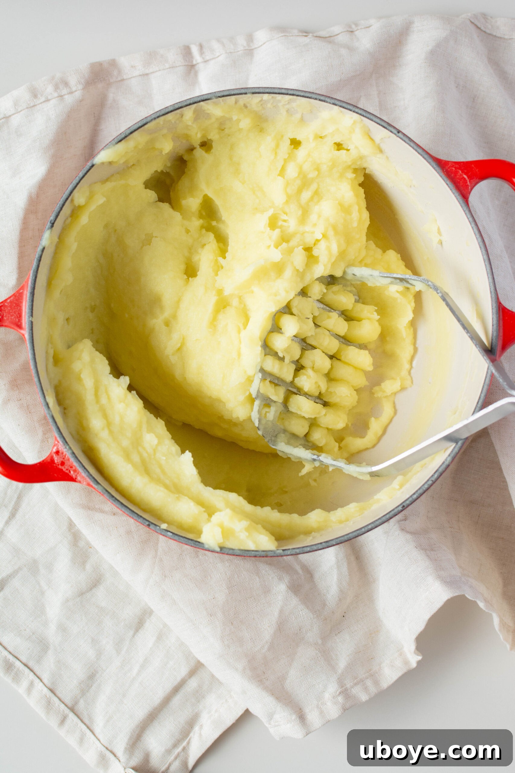 Large red pot filled with olive oil mashed potatoes, with a potato masher resting inside.