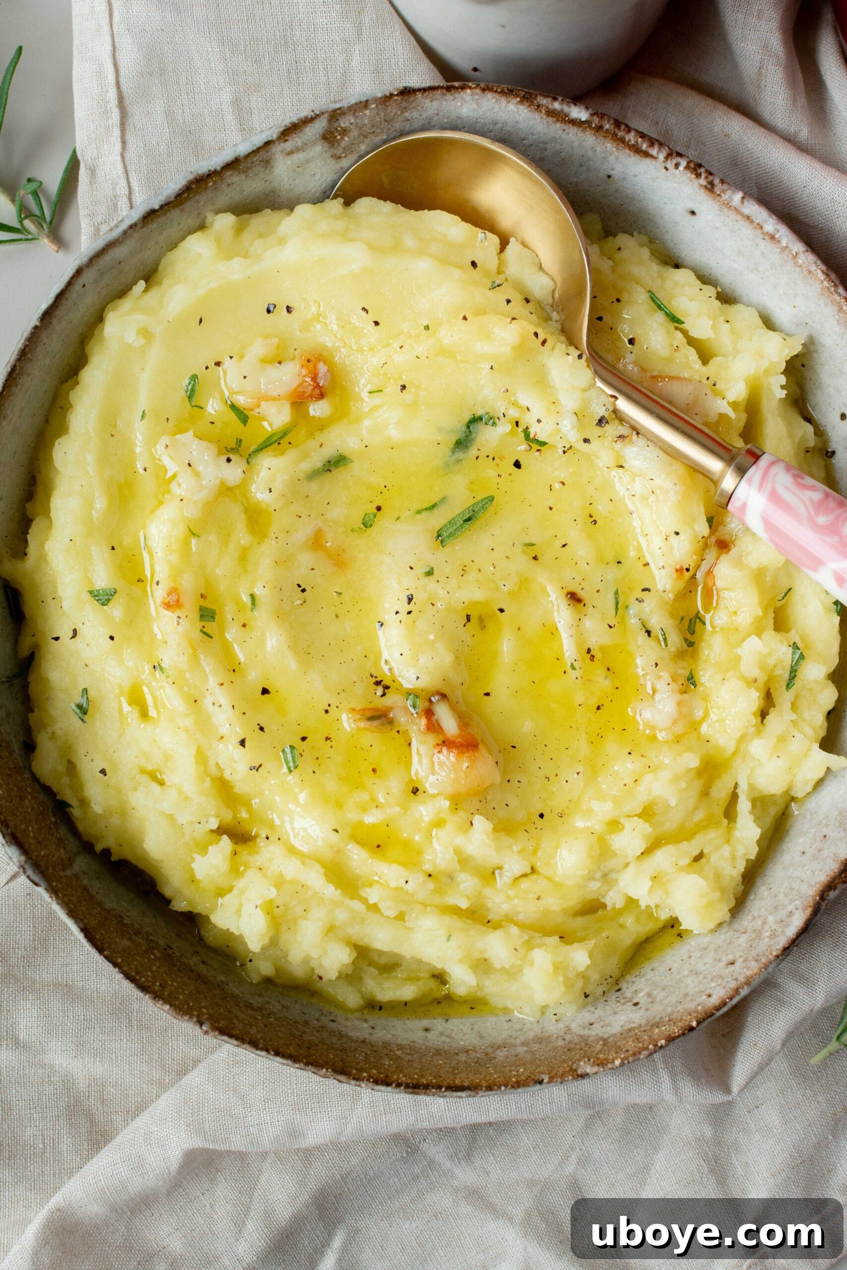 Bowl of olive oil mashed potatoes garnished with fresh rosemary and black pepper, with a gold spoon.
