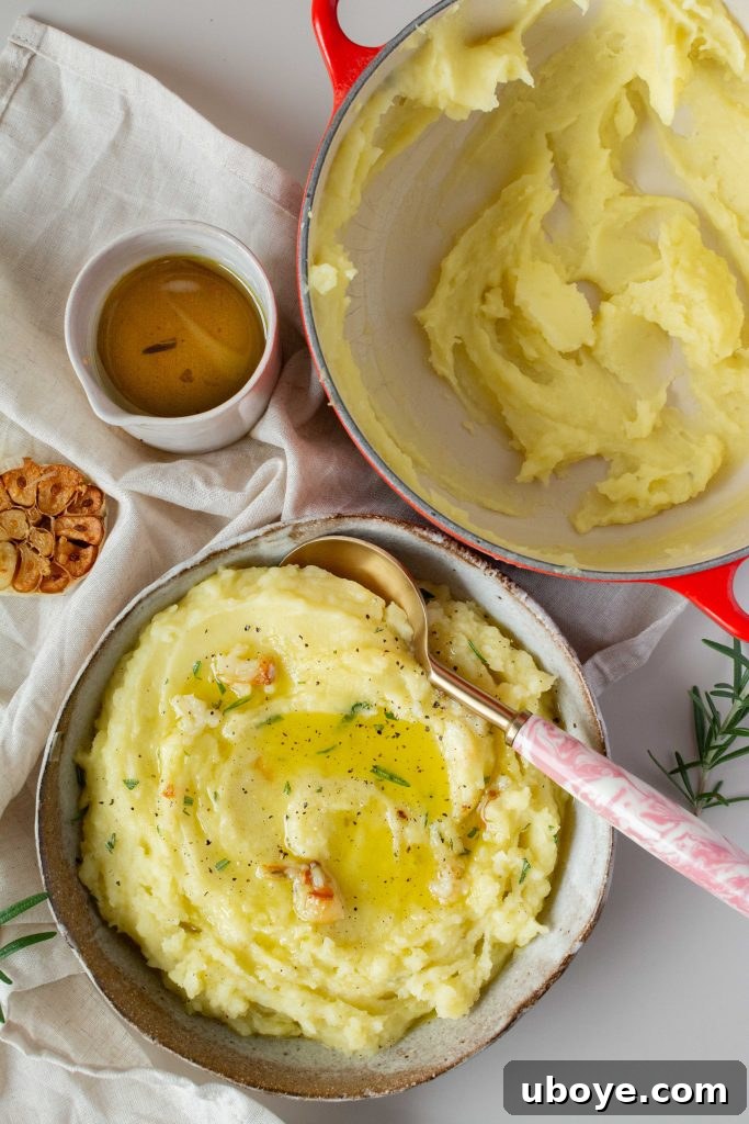 A bowl of olive oil mashed potatoes with rosemary, a spoon, and a red pot of mashed potatoes in the background.