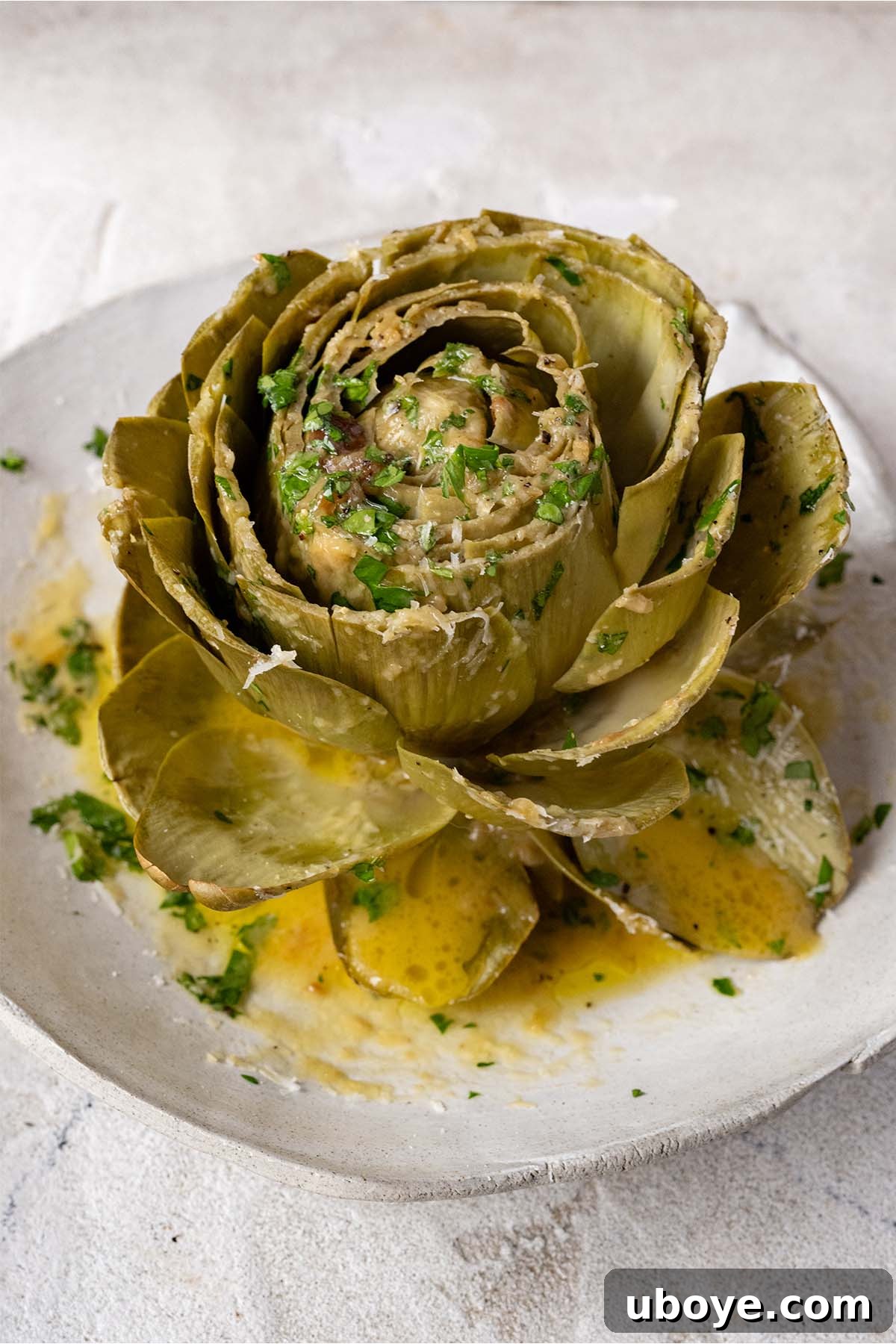 A beautifully presented large steamed artichoke, garnished with melted garlic confit butter, grated Parmesan cheese, and fresh herbs on a white plate.