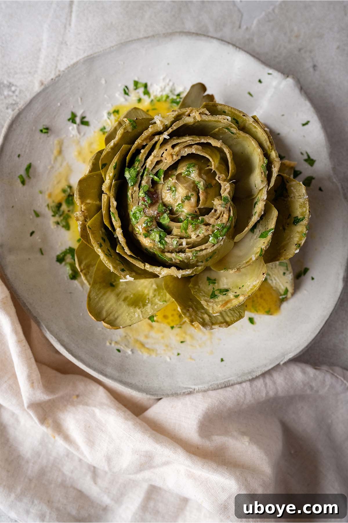 A whole steamed artichoke adorned with generous amounts of melted garlic confit butter, grated Parmesan, and fresh parsley, ready to be enjoyed.