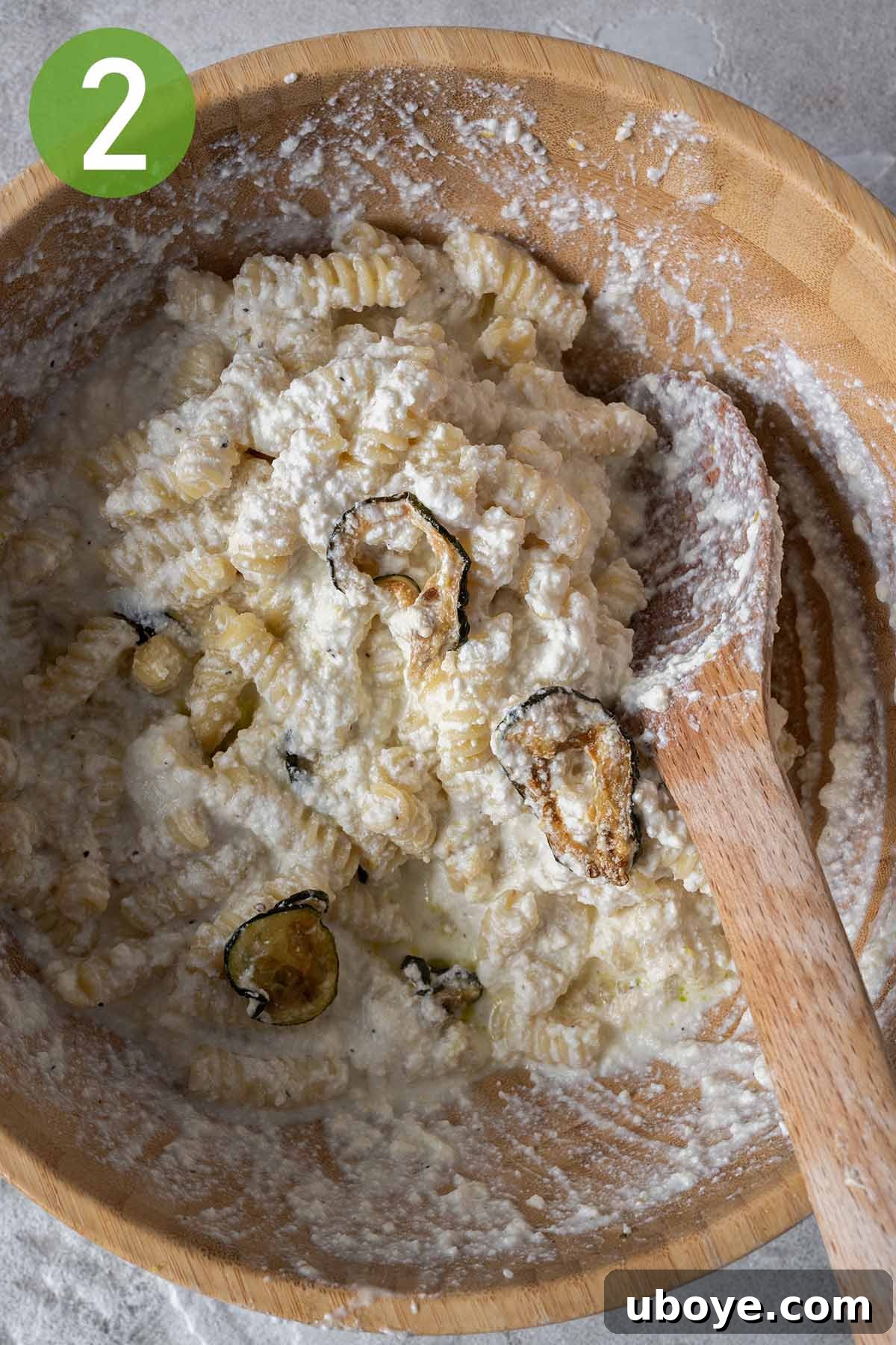 Cooked pasta and crispy fried zucchini being gently mixed into a bowl of creamy lemon ricotta sauce, ensuring every strand is coated.