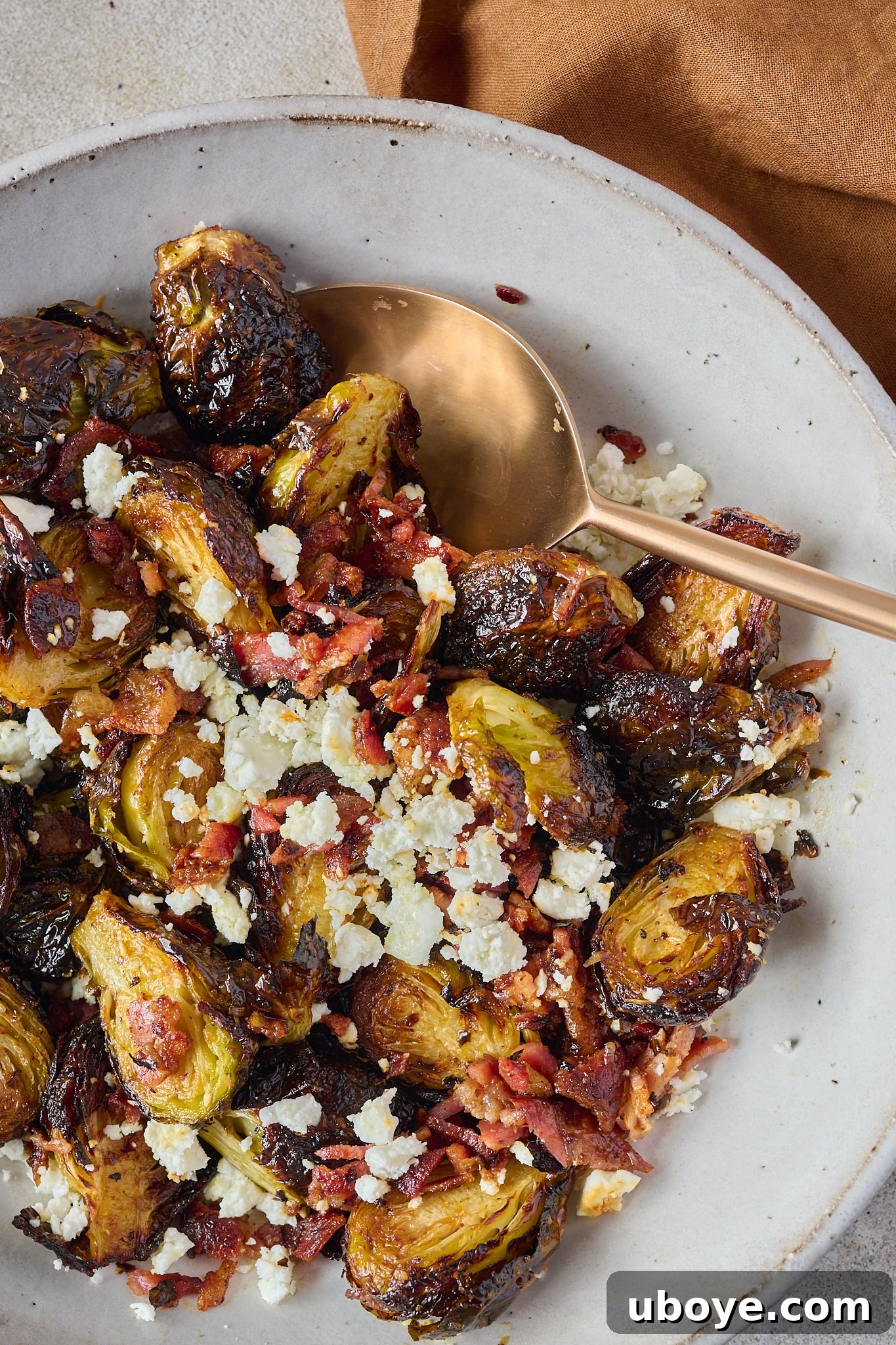 Crispy Brussels sprouts with bacon and crumbled feta in a large white bowl, with a gold spoon.