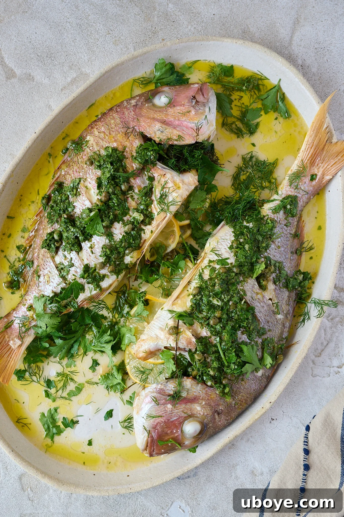 A beautifully roasted whole baked snapper, generously garnished with fresh herbs and a drizzle of salsa verde, rests on a large white platter, ready for serving.